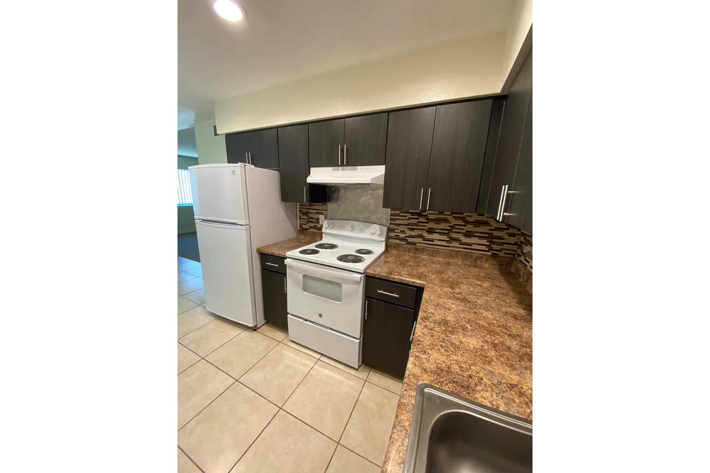 A modern kitchen featuring dark cabinetry, a white stove and range hood, and a white refrigerator. The countertops are a warm brown with a tiled backsplash consisting of small, multicolored tiles. The space has neutral tile flooring and ample natural light from a window in the background.