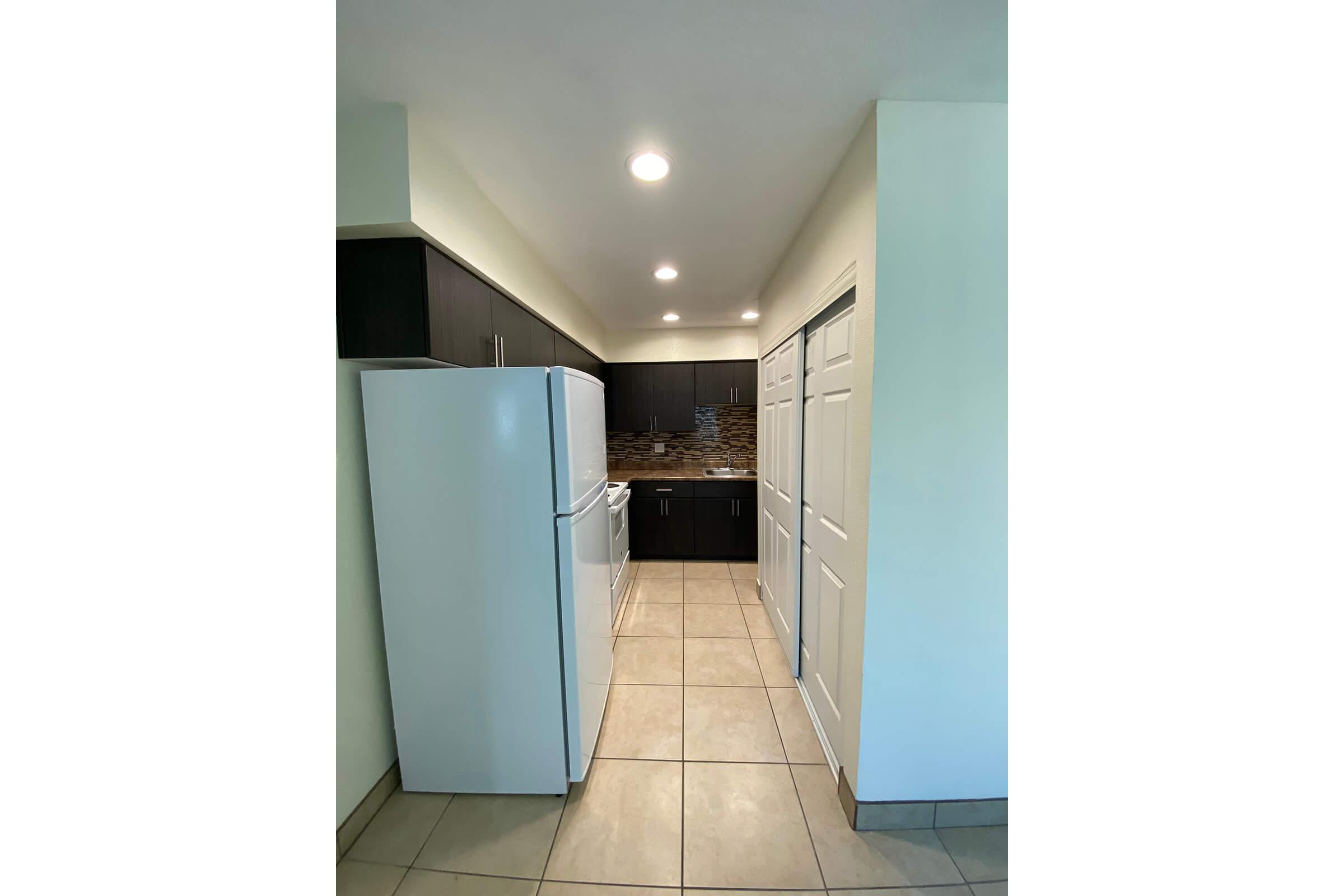 A narrow kitchen featuring a white refrigerator and oven, set against dark cabinetry. The walls are light-colored, and the floor has beige tiles. A row of cabinets is visible on one side, with double doors leading to a storage area. Bright overhead lights illuminate the space, creating a clean and modern look.