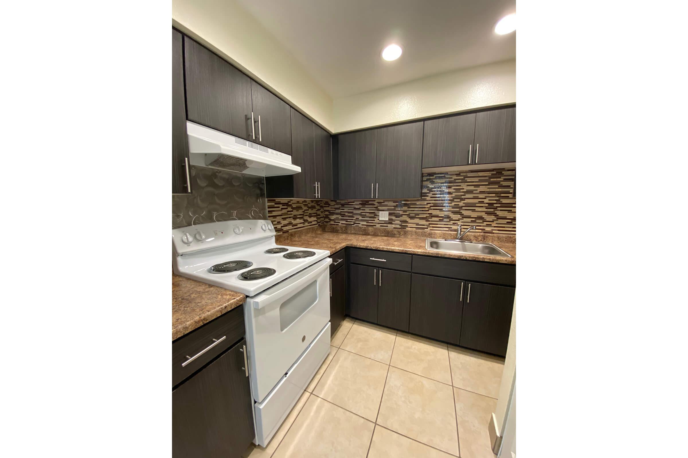 A modern kitchen featuring dark wooden cabinets, a white oven and stovetop, a stainless steel sink, and a tiled backsplash with brown and beige tones. The countertop is a light brown material, and the floor is tiled in a neutral color. Bright overhead lighting illuminates the space.