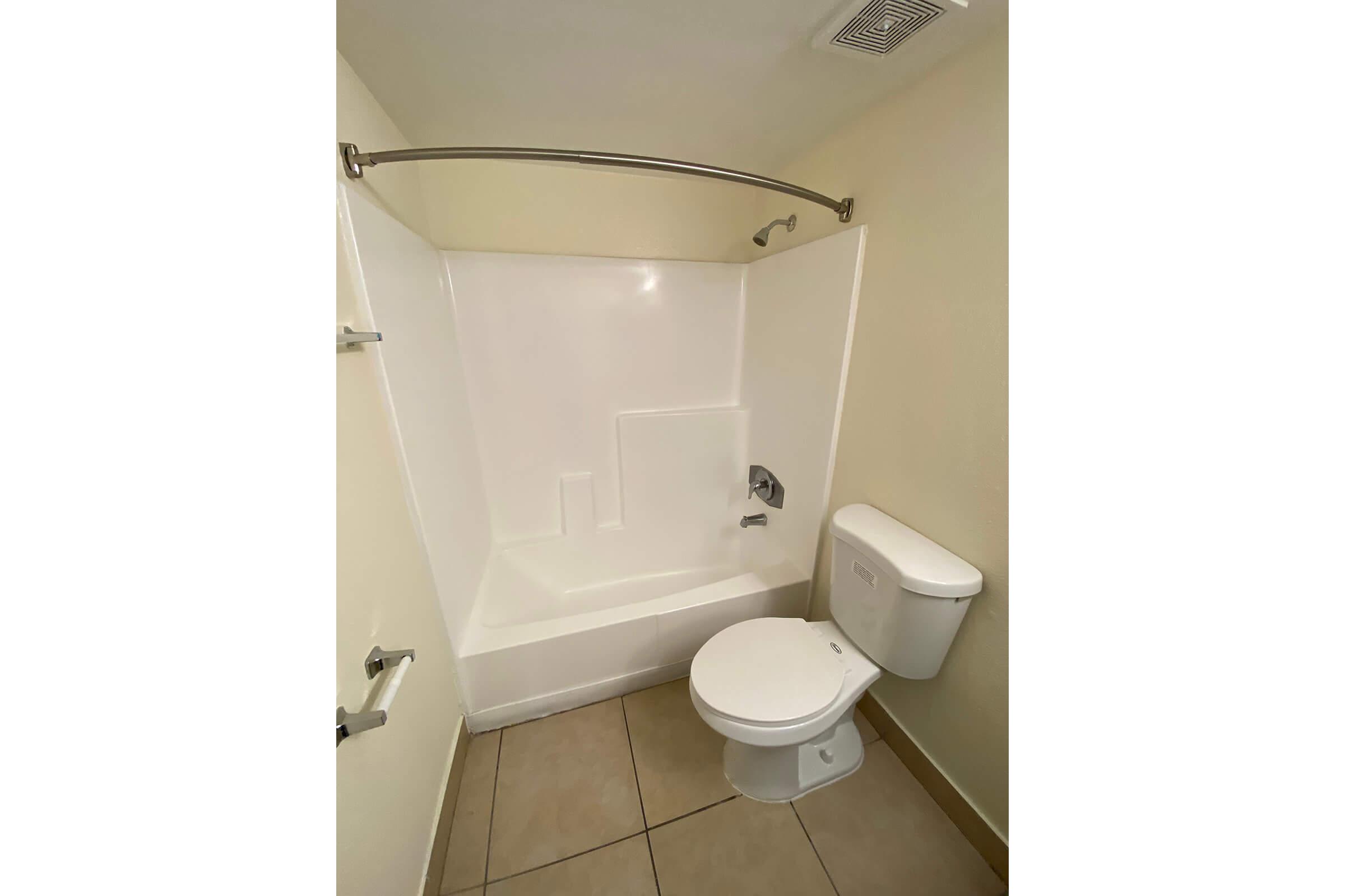 A clean, simple bathroom featuring a white bathtub with a shower curtain rod, a modern toilet, and beige walls. The floor is tiled, and the overall look emphasizes minimalism and functionality.