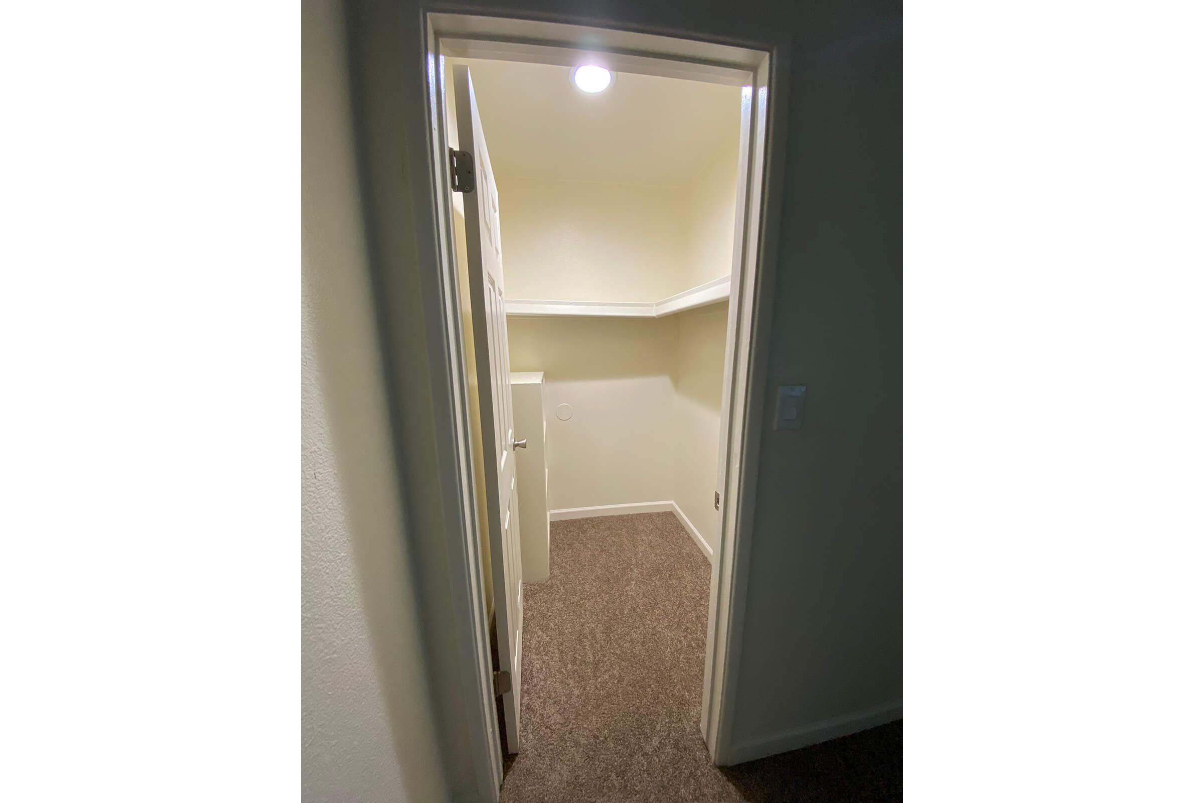 A view of a small, partially enclosed room with beige walls and a light overhead. The entrance shows a door leading into the space, which has carpeted flooring and a built-in shelf along one wall, creating a tidy and organized appearance. A light switch is visible to the right of the doorway.