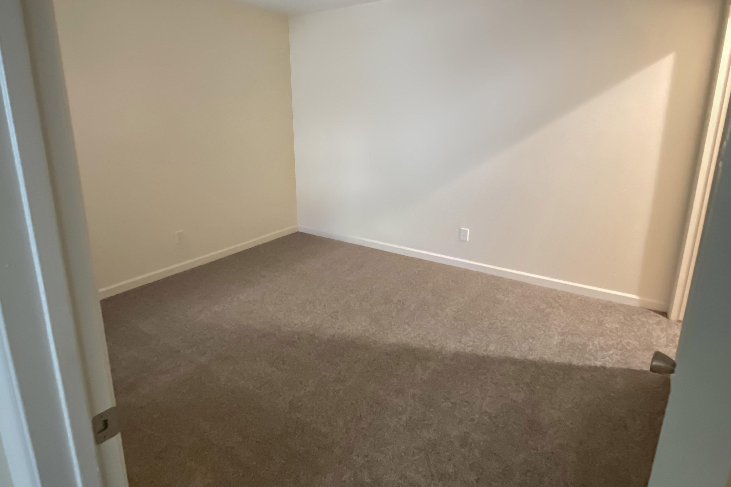 Empty room with light beige walls and gray carpet. The space has no furniture and is well-lit, with a soft shadow across the floor. A closed door is partially visible on the left side. The overall atmosphere is clean and minimalist.