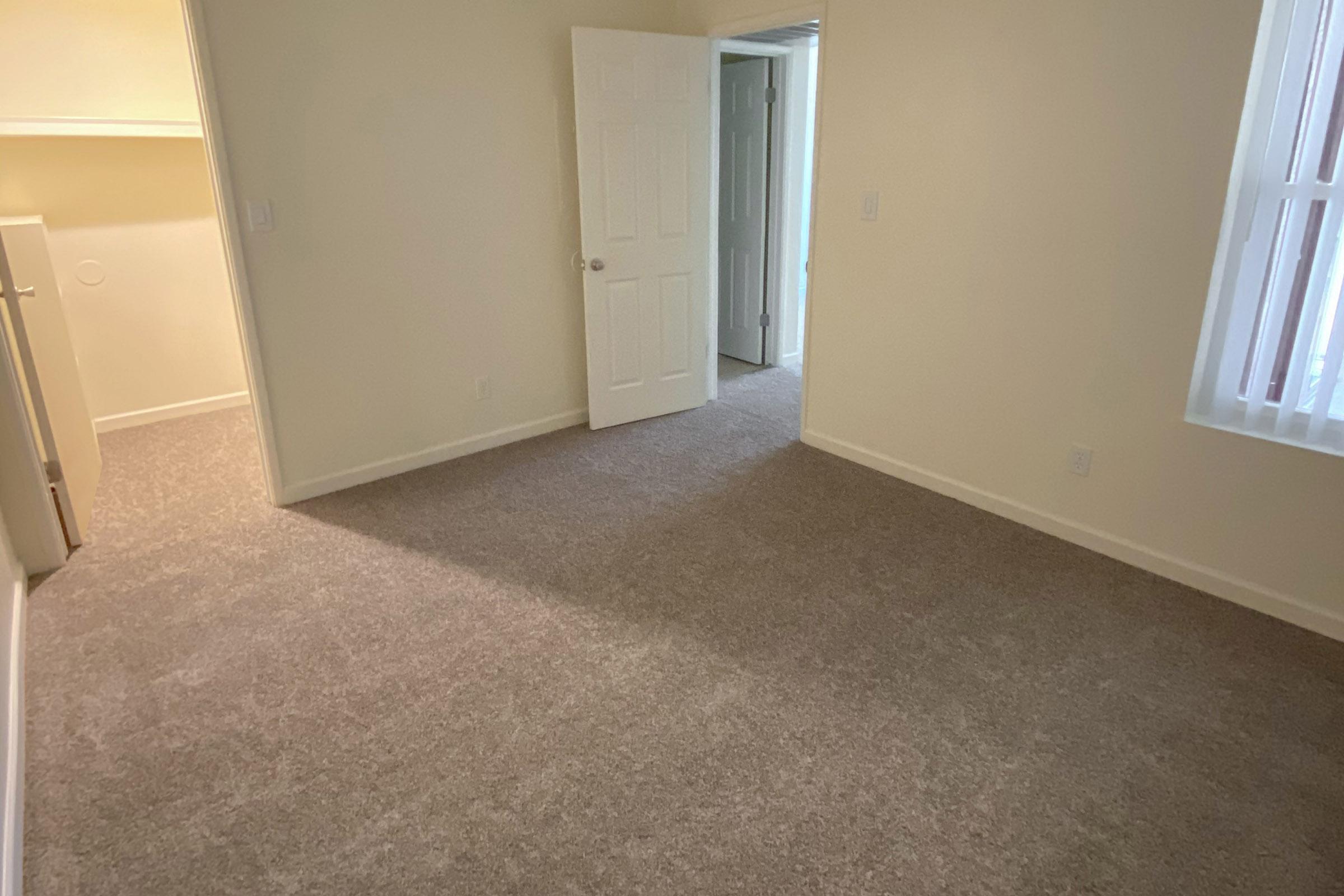 A vacant room with light beige walls and freshly carpeted flooring. There is a partially open door leading to another room, and a closet can be seen on the left side. Natural light filters through a window, creating a bright and airy atmosphere in the space.