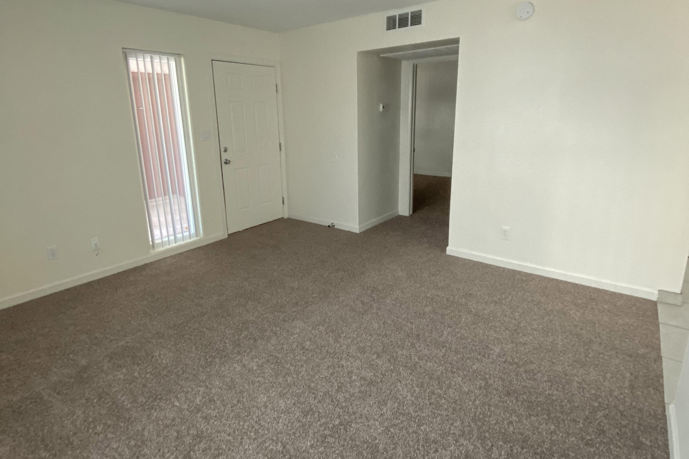 A vacant room with beige carpet and light-colored walls. A door leads outside, and a window is visible on the left side. There is a small hallway to the right that leads to another room, emphasizing an open and spacious layout.