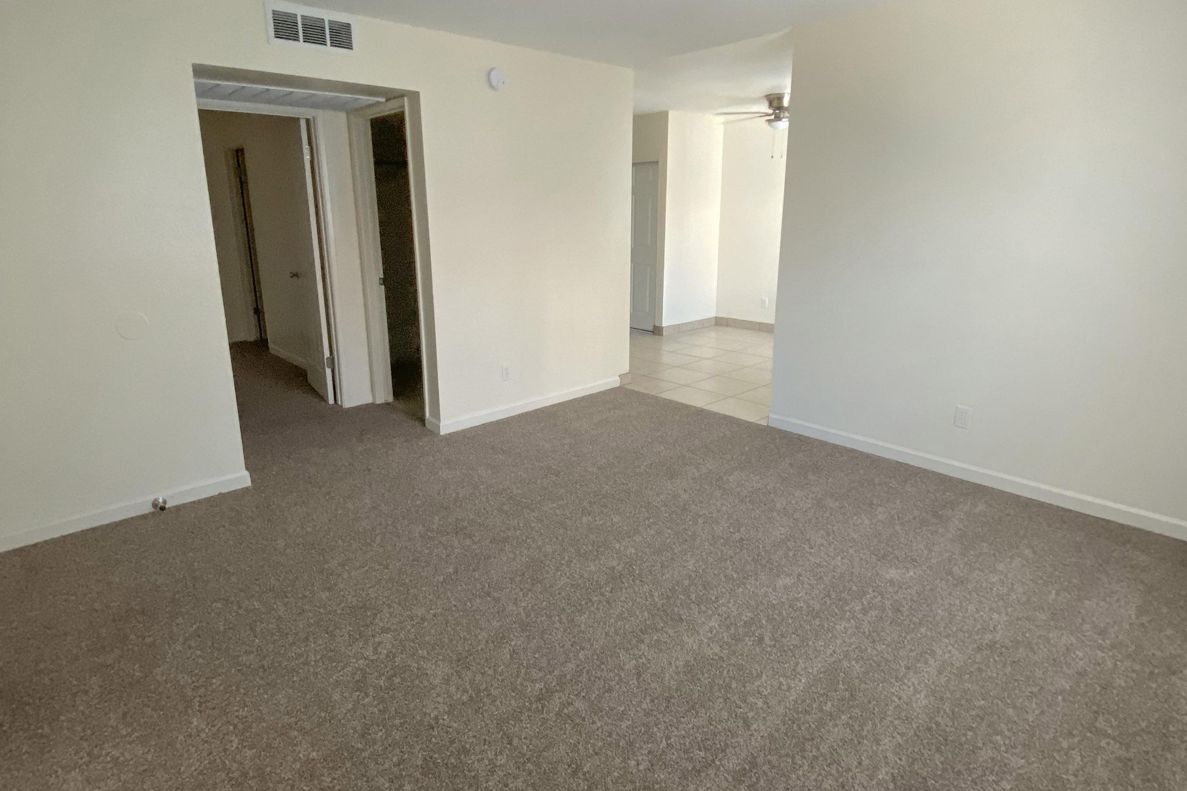 A vacant room with light-colored walls and beige carpet. There is a doorway leading to another room on the left, and a tile floor visible in the background. The lighting is bright, creating an open and spacious feel. A ceiling fan is visible in the upper right corner.