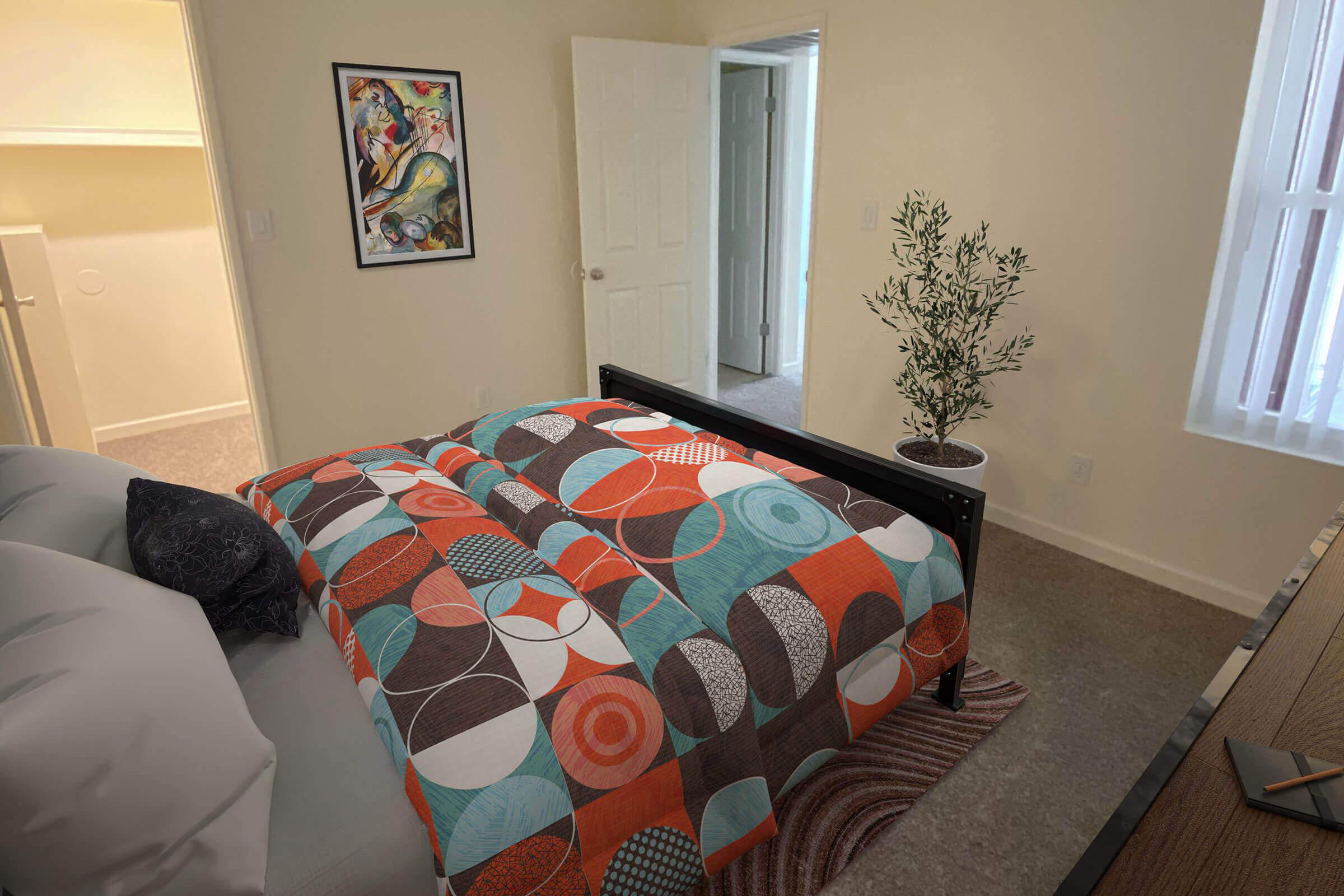 A cozy bedroom featuring a bed with a colorful patterned duvet in shades of orange, blue, and white. There is a small potted plant beside the bed, a rug on the floor, and a framed artwork on the wall. An open door leads to another room, and natural light fills the space through a window.