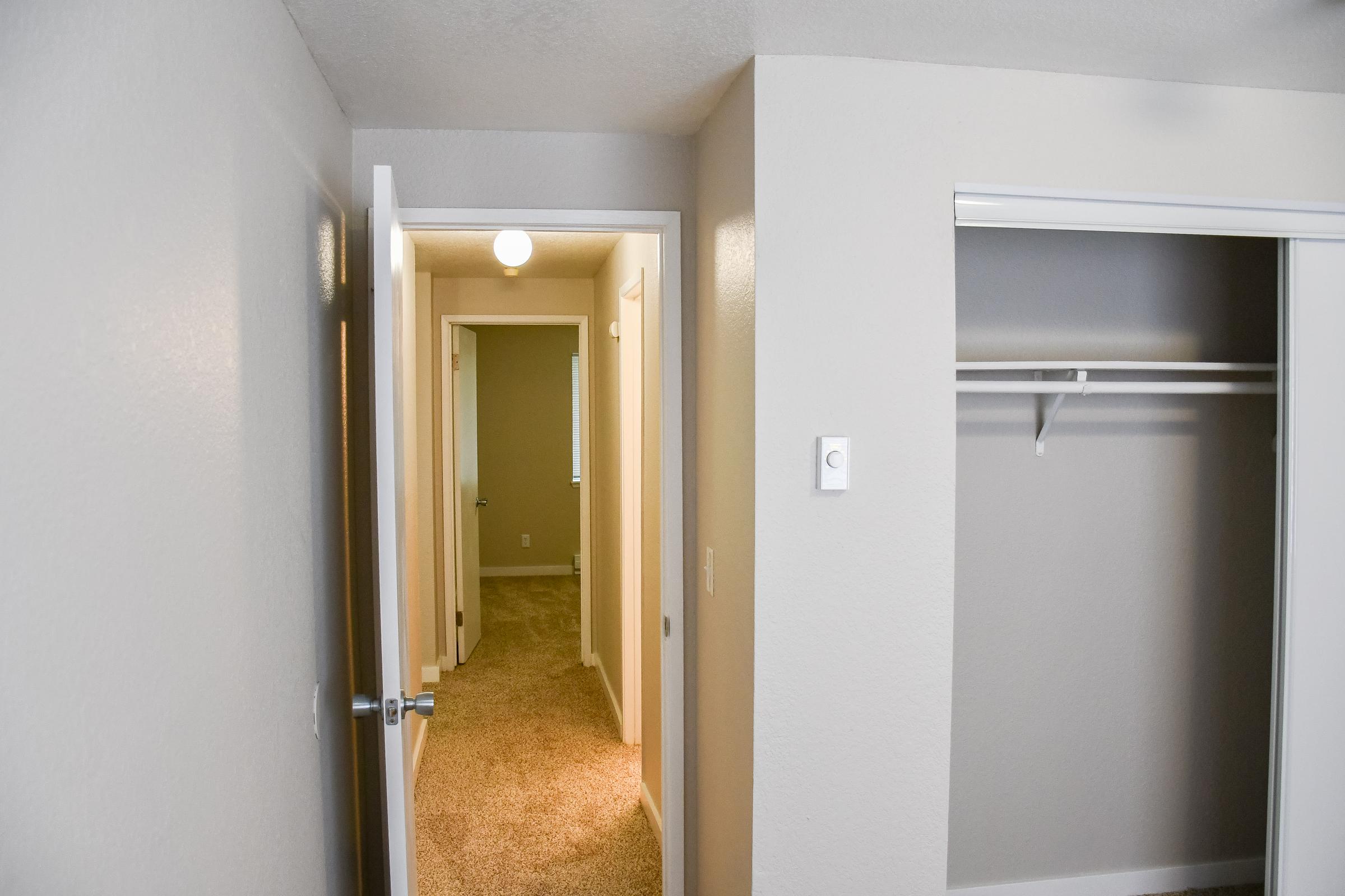 A view of an interior space featuring a light-colored, carpeted hallway with doors leading to other rooms. The walls are painted in neutral tones, and there is an open closet with a single shelf on the left side. Soft overhead lighting creates a bright atmosphere.