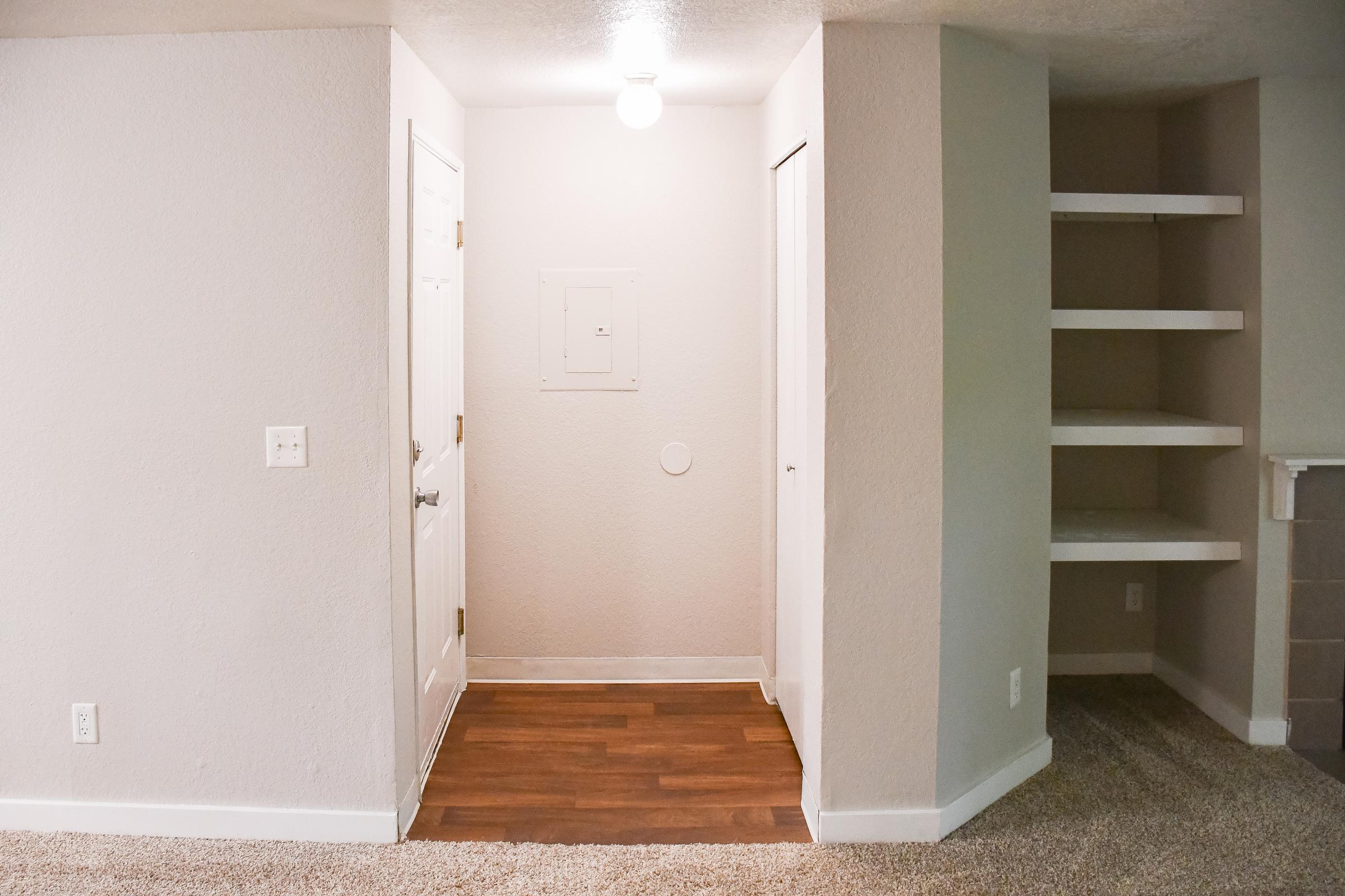 A bright, neutral-colored entrance area featuring a light fixture and a wall-mounted electrical panel. There are two closed doors, one leading into a closet and another possibly leading to a room. To the right, a built-in shelving unit is visible against the wall, with a soft carpeted floor beneath.