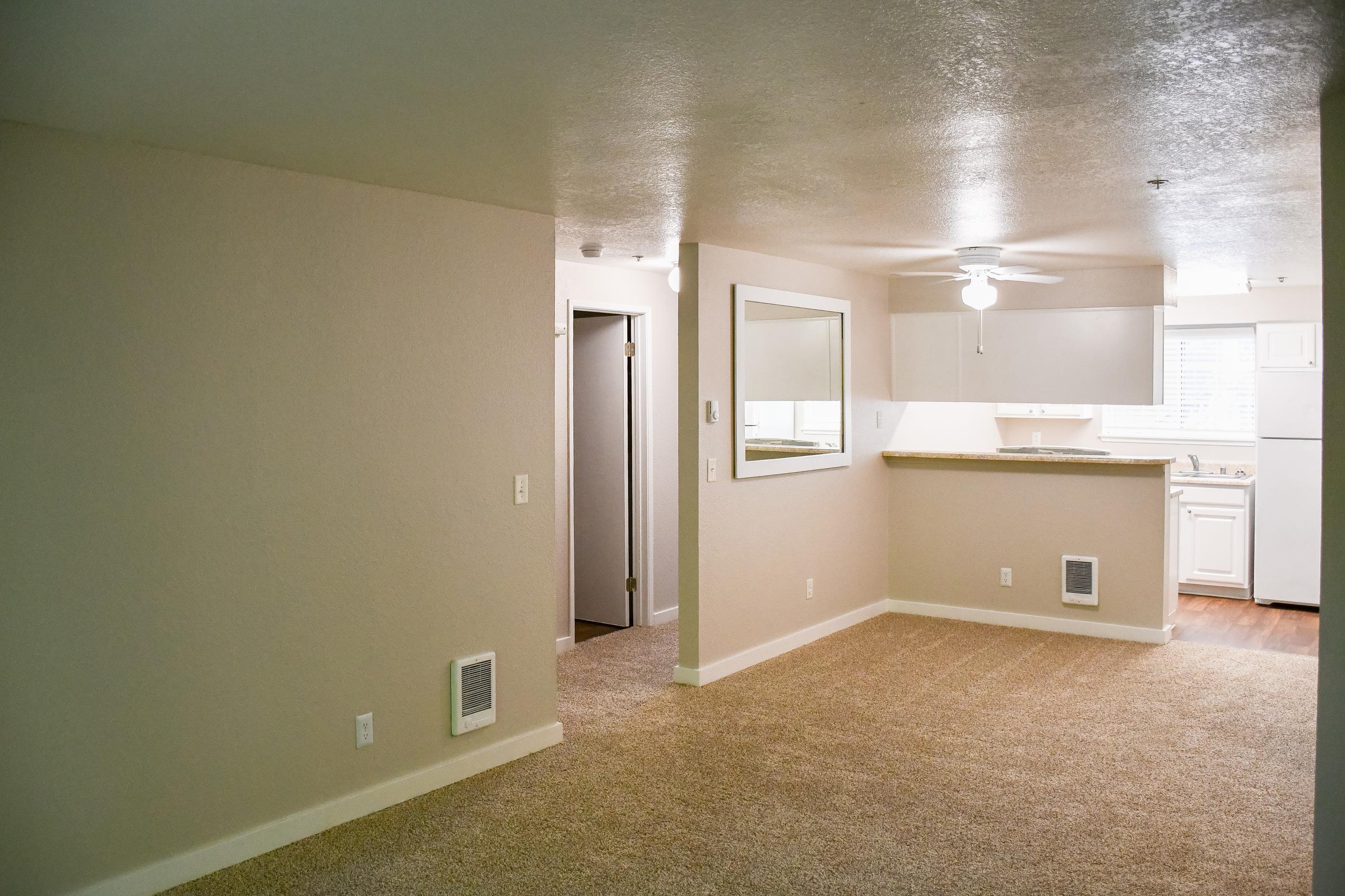 A well-lit, spacious room featuring beige walls and carpet, with an open layout leading to a kitchen. A ceiling fan is visible, along with a mirrored wall. A doorway leads to another room, and there are vents on the walls, highlighting the modern design of the space. Natural light streams in through a window.