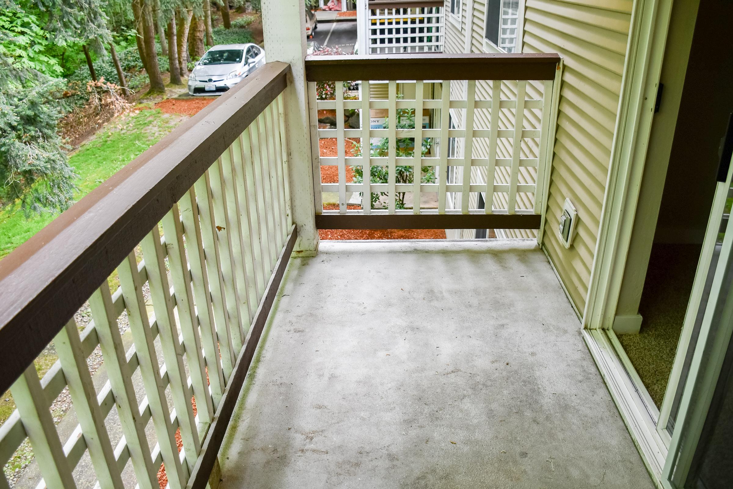 A small concrete balcony with a wooden railing, overlooking a green yard and trees. A parked white car is visible in the background, along with another building. The balcony is empty, with a clean surface and no furniture. Natural light illuminates the area.