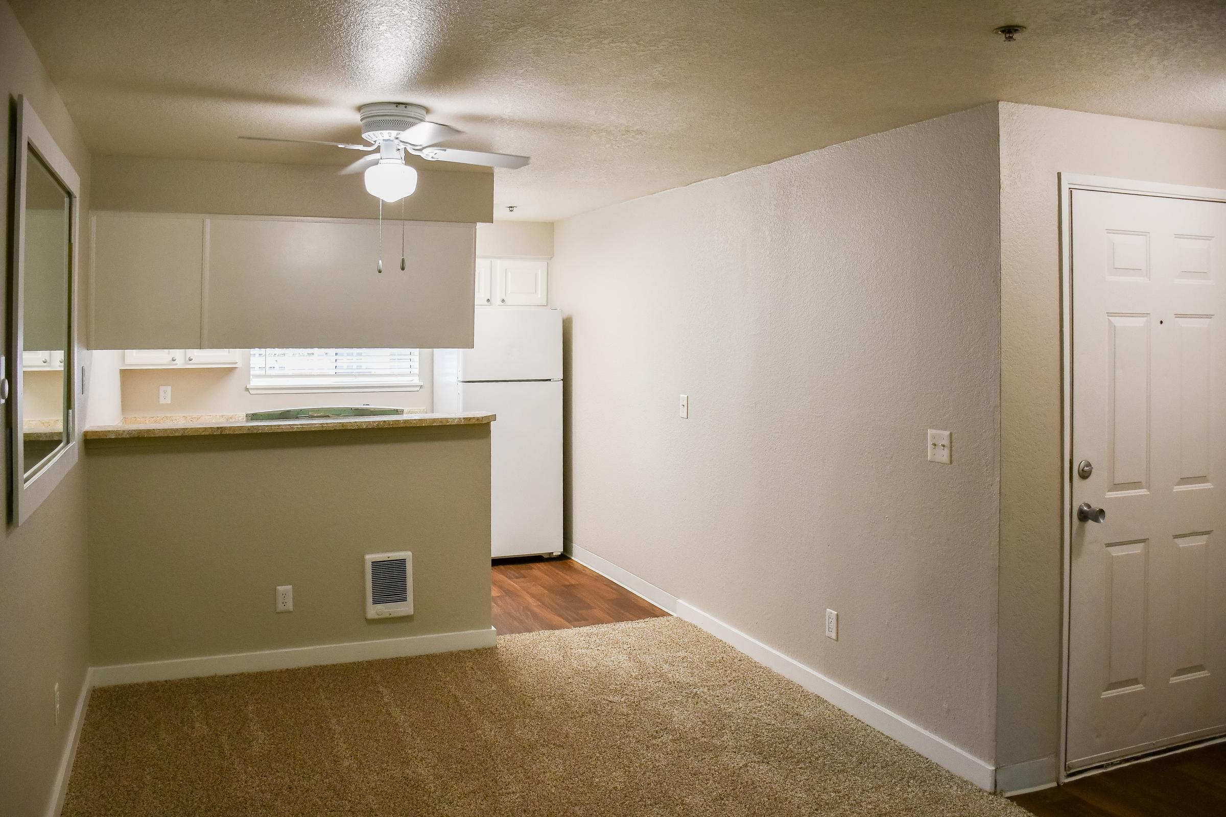 An interior view of a small apartment showcasing a living area with carpeted flooring, a ceiling fan, and a doorway leading to the outside. The kitchen is partially visible with white cabinetry and a refrigerator. Natural light enters through a window near the kitchen area.