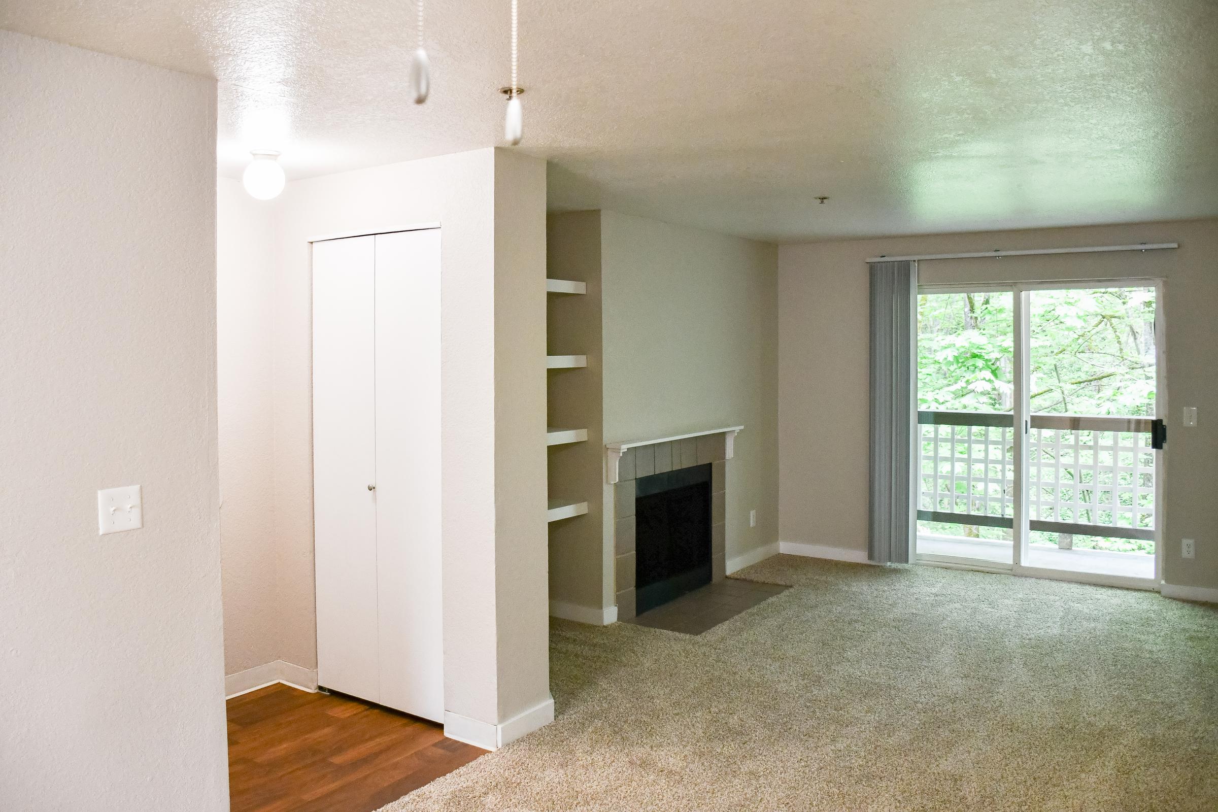 A bright, spacious living area featuring beige walls, a light carpeted floor, and a white fireplace. There's a door leading to a balcony with greenery visible outside. A closet is positioned to the left, and shelving is integrated into the wall beside the fireplace. Natural light floods the room from large sliding glass doors.