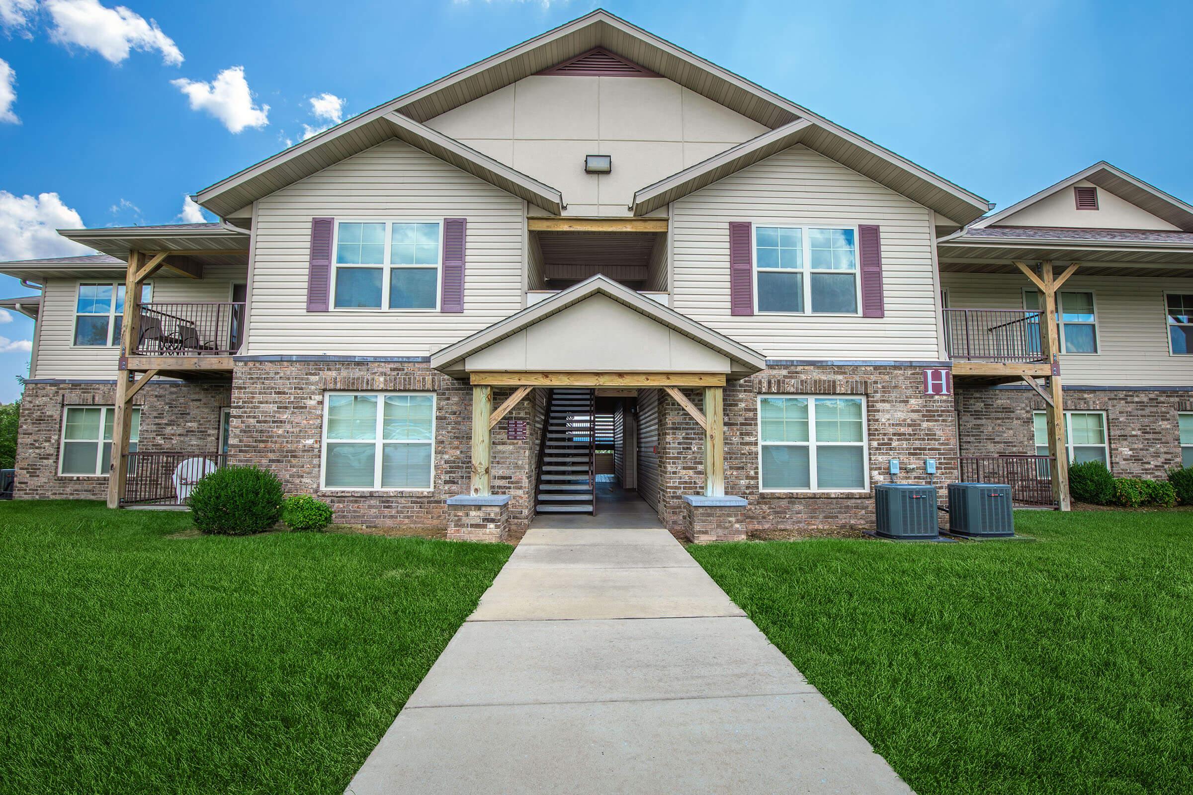 A two-story apartment building featuring a brick and siding exterior. The entrance is flanked by large windows and a wooden awning above the door. A well-maintained pathway leads to the entrance, with green grass surrounding the building and two air conditioning units visible on the side. Scattered clouds are visible in the blue sky.