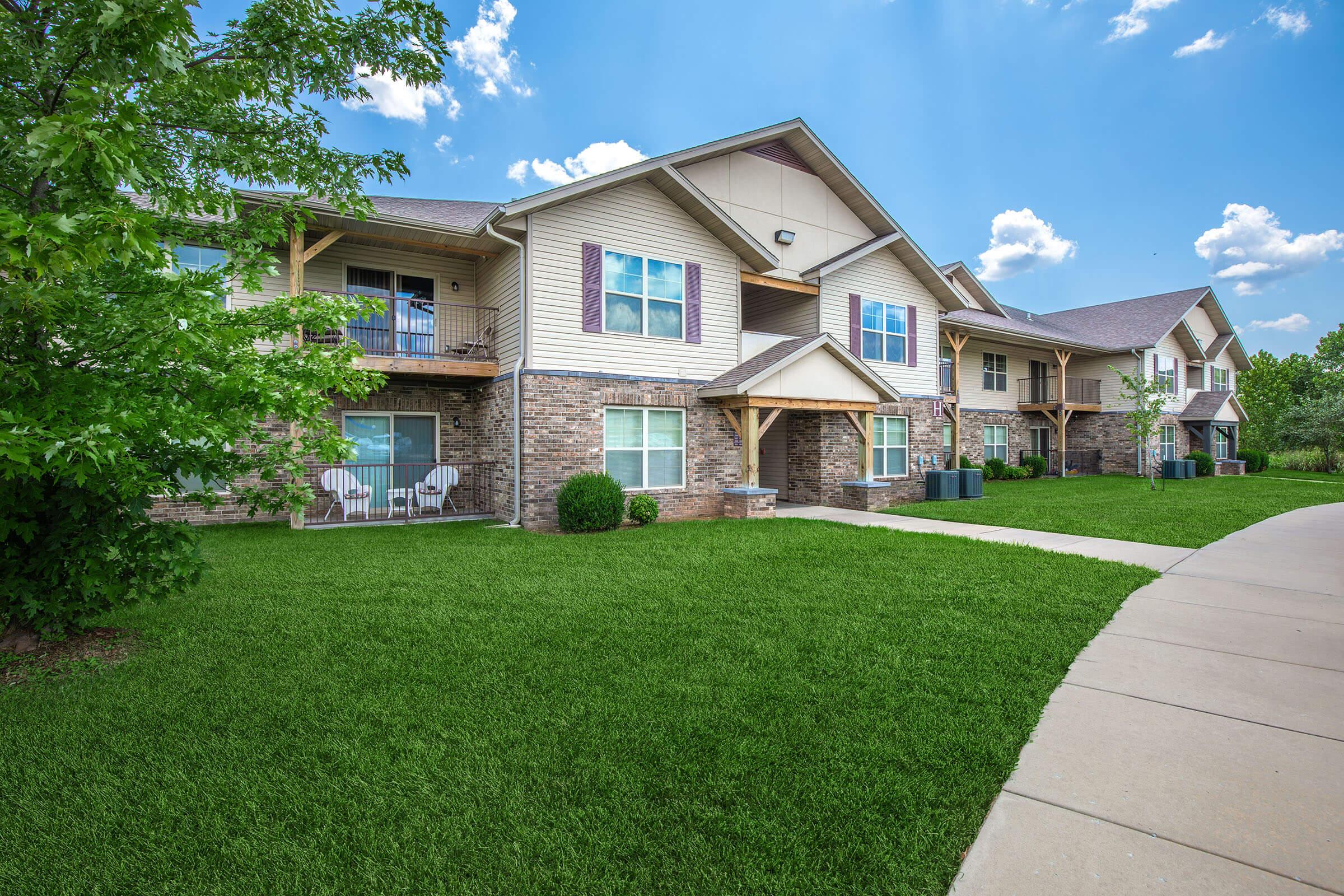 A row of modern apartment buildings with beige siding and stone accents, surrounded by well-maintained lawns and landscaping. The sky is bright blue with fluffy white clouds, and there are chairs on the balconies of the apartments. A curved sidewalk runs in front of the buildings, indicating a well-manicured community.