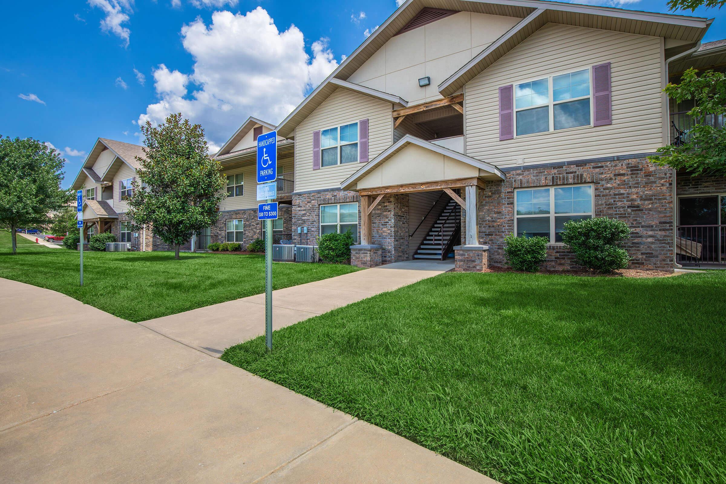 A residential building with a stone and siding facade, featuring staircases and accessible parking signs. The surrounding area has lush green lawns and clear blue skies with a few clouds. Sidewalks lead to the entrance.