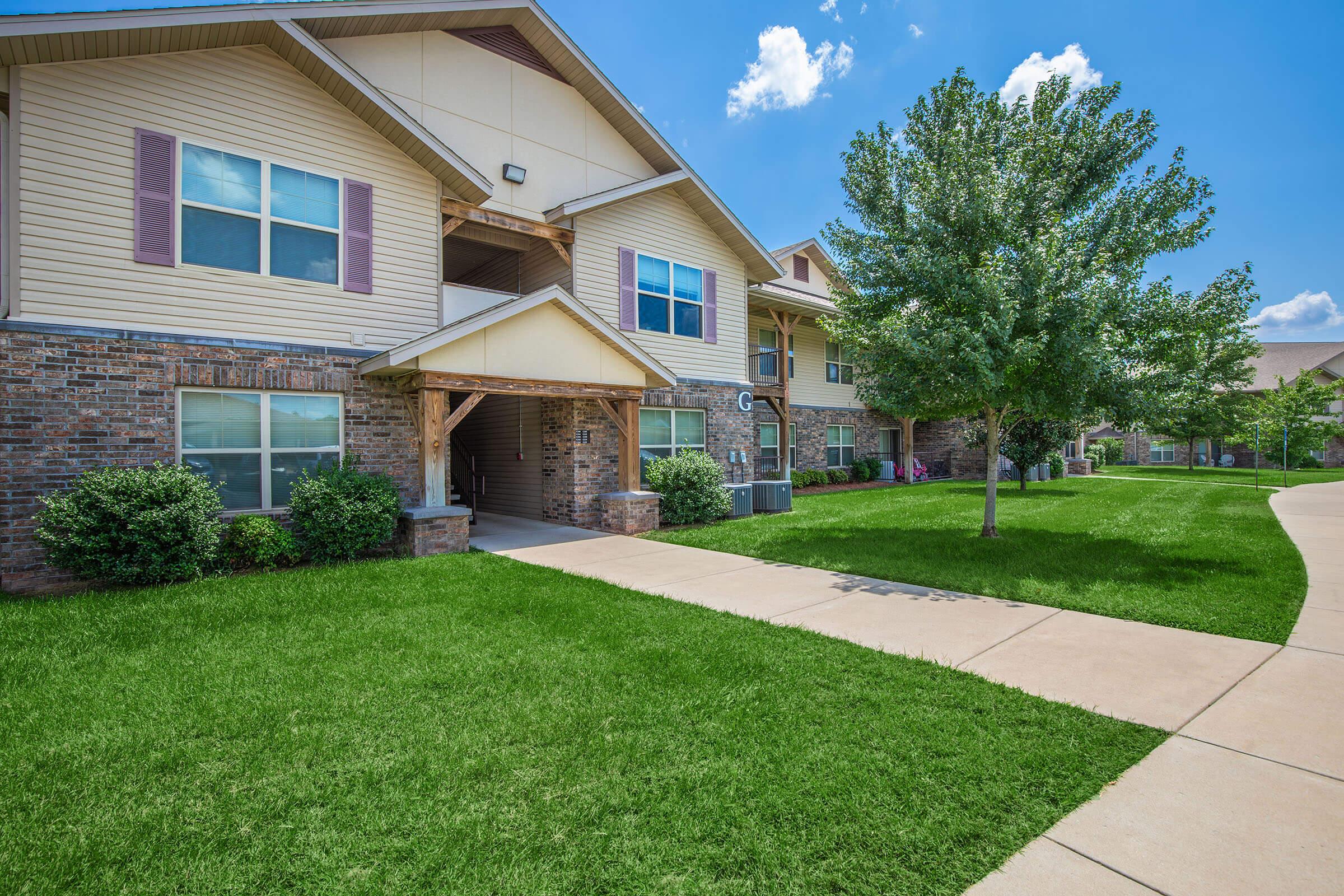 A well-maintained apartment complex showcasing two-story buildings with beige siding and brick accents. Green lawns and trees surround the walkway, under a clear blue sky with fluffy white clouds. The scene conveys a clean and inviting outdoor environment.