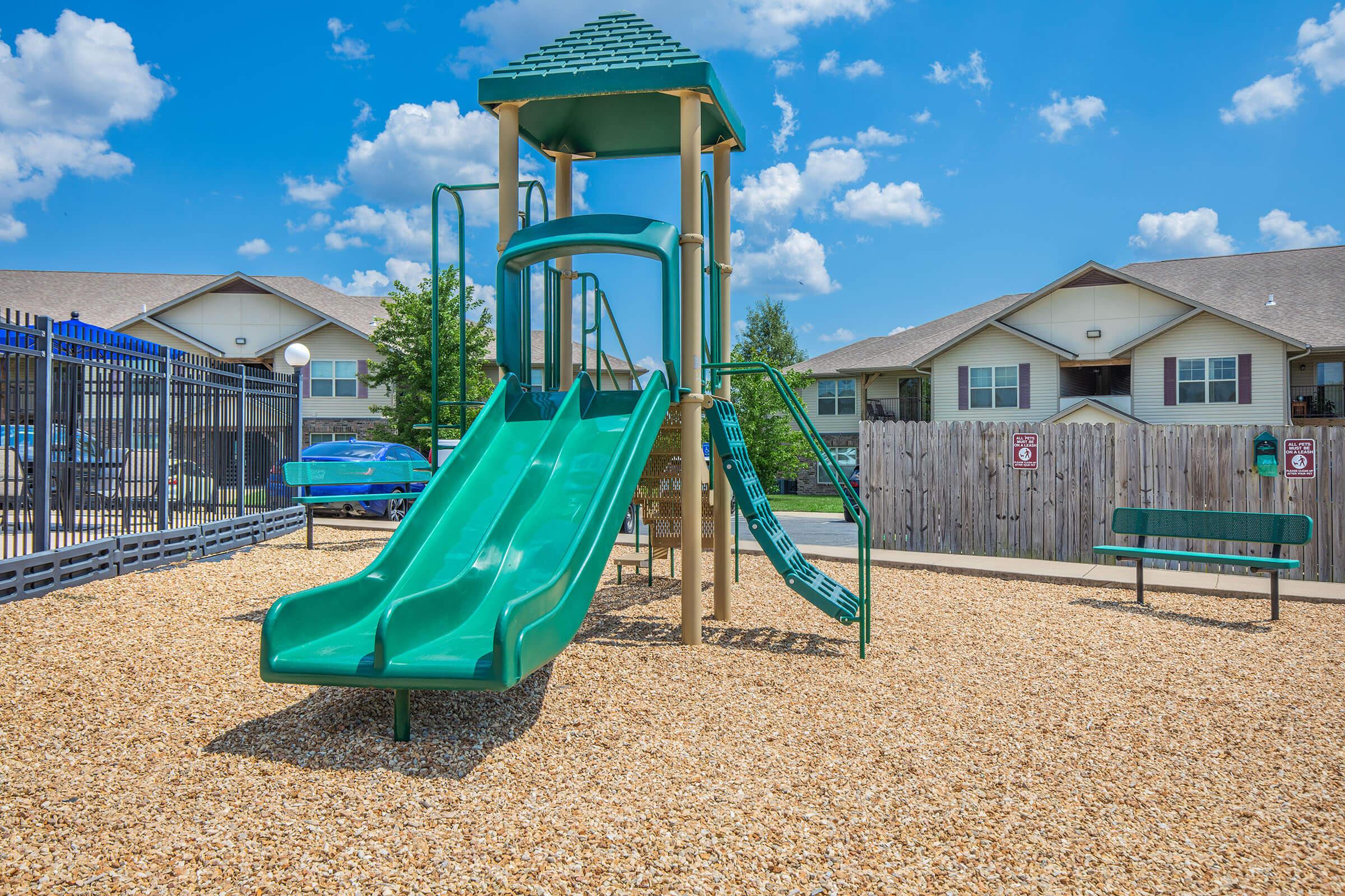 A green playground with a slide, climbing structures, and a sandbox, surrounded by gravel. In the background, there are residential buildings and a fence. The sky is bright blue with a few fluffy clouds, and a bench is visible nearby.