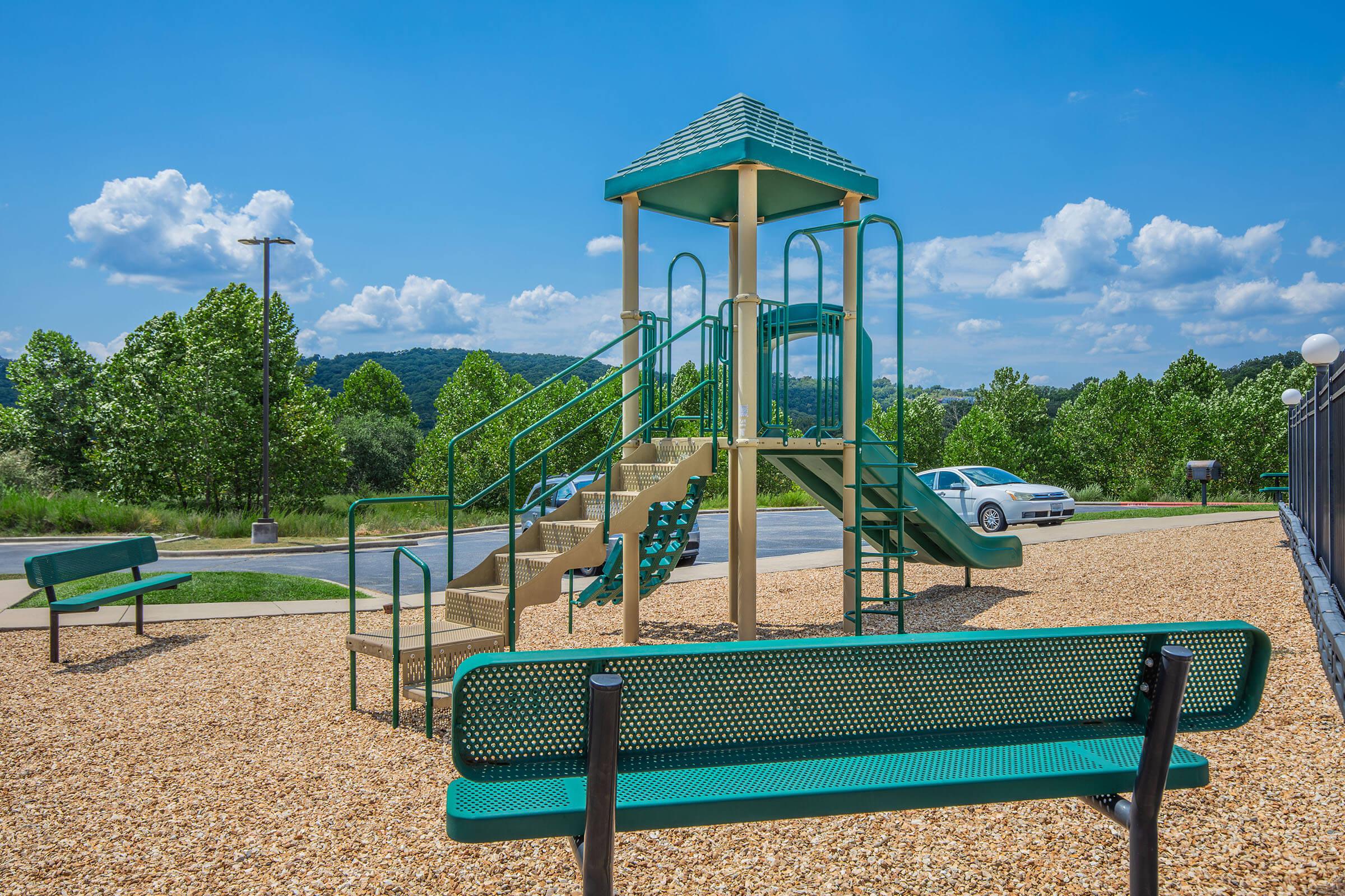 A green and beige playground with a multi-level slide and stairs, surrounded by a gravel surface. In the background, there are trees and a clear blue sky with scattered clouds. Two green benches are placed nearby, and a parked white car is visible on the road.