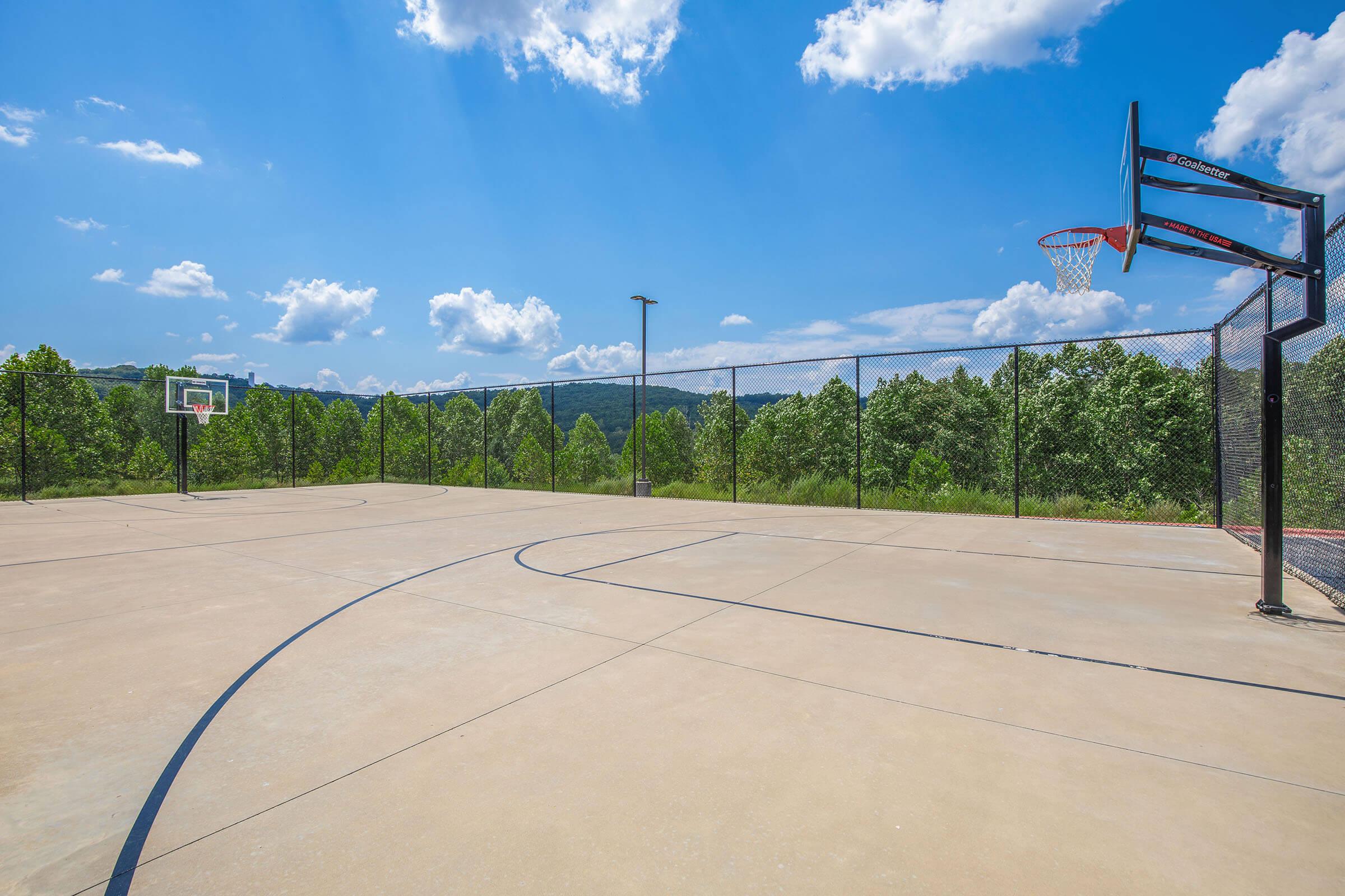 A bright outdoor basketball court with two hoops, surrounded by greenery. The court has a clear blue sky above with fluffy white clouds. It's empty and features a smooth concrete surface, providing an open space for basketball activities.