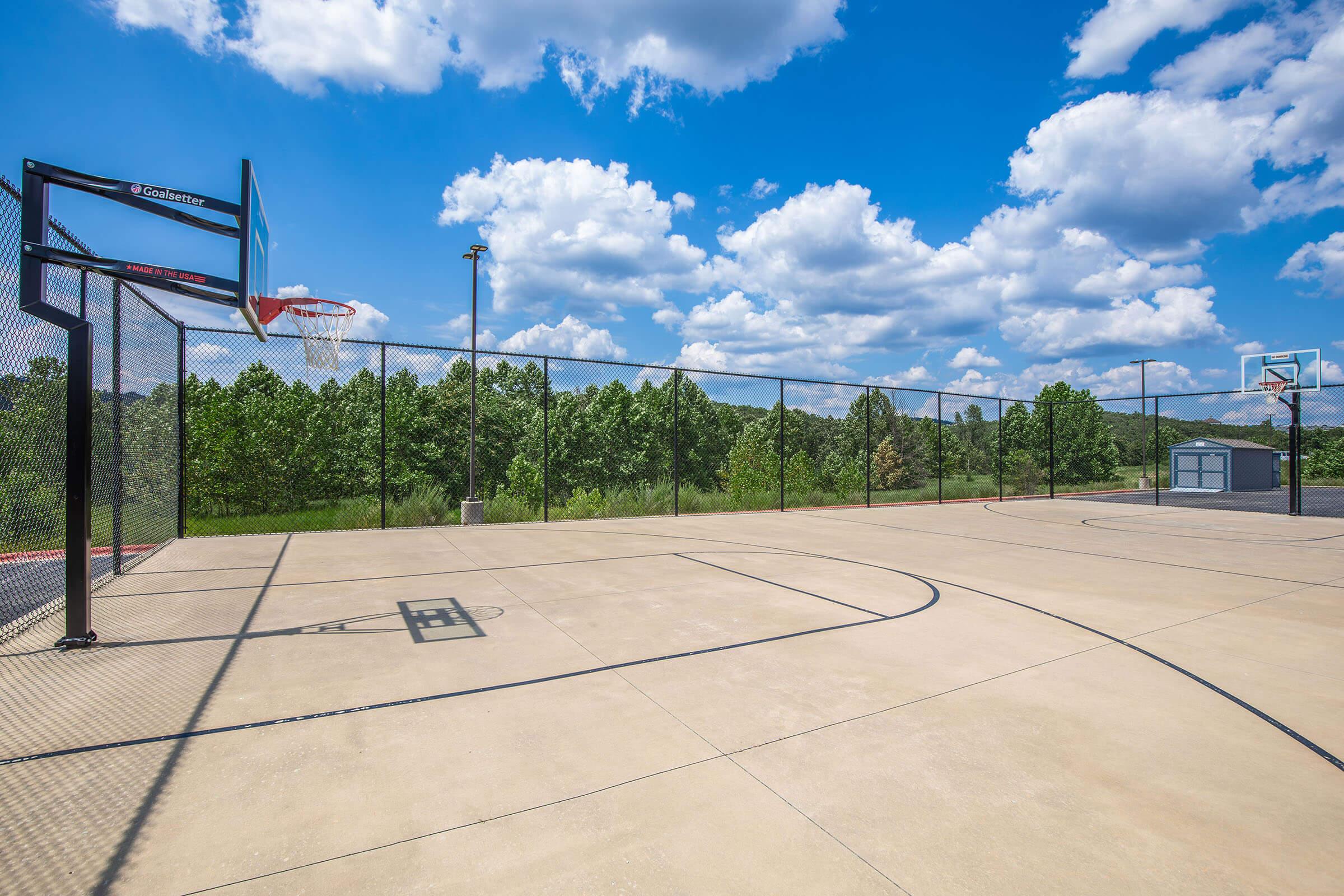 A sunny basketball court featuring two hoops, surrounded by a fence. The smooth concrete surface shows clear markings for gameplay, with lush green trees and a blue sky filled with fluffy white clouds in the background.