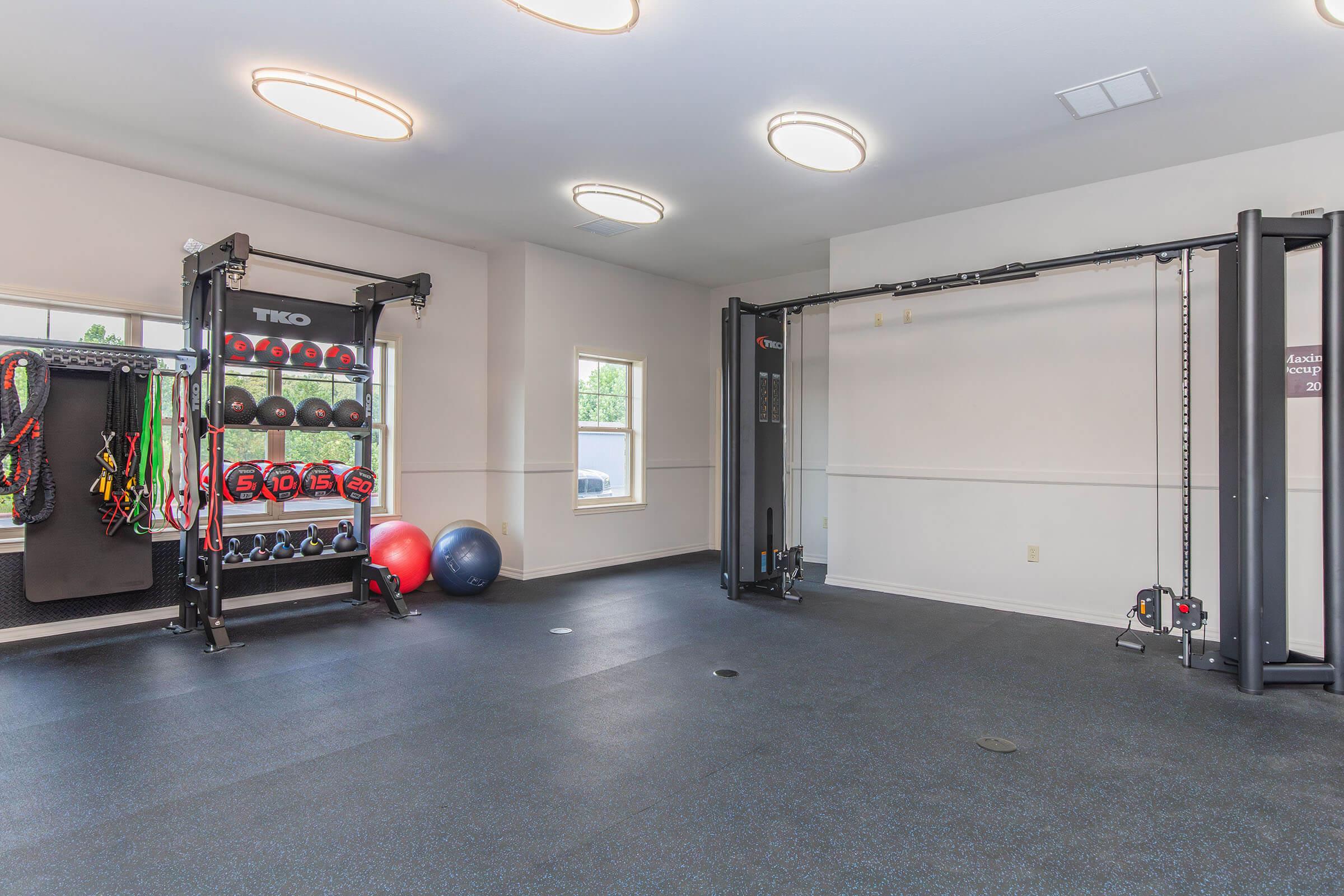 A modern gym interior featuring a cable machine, weight storage rack filled with various weights, exercise balls, and resistance bands. The flooring is dark and the walls are light-colored, creating a spacious environment. Overhead lighting provides bright illumination in the workout space.