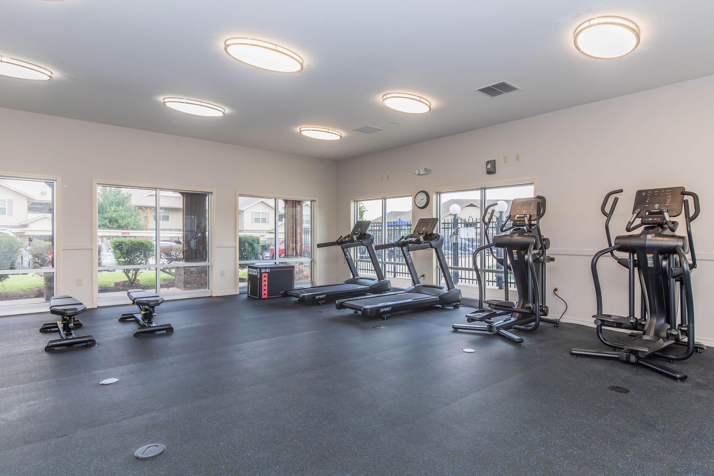 A spacious gym featuring multiple treadmills and elliptical machines, along with a weight bench. Large windows allow natural light to fill the area, offering a view outside. The flooring is dark and rubberized, suitable for workouts. Ceiling lights provide additional illumination.