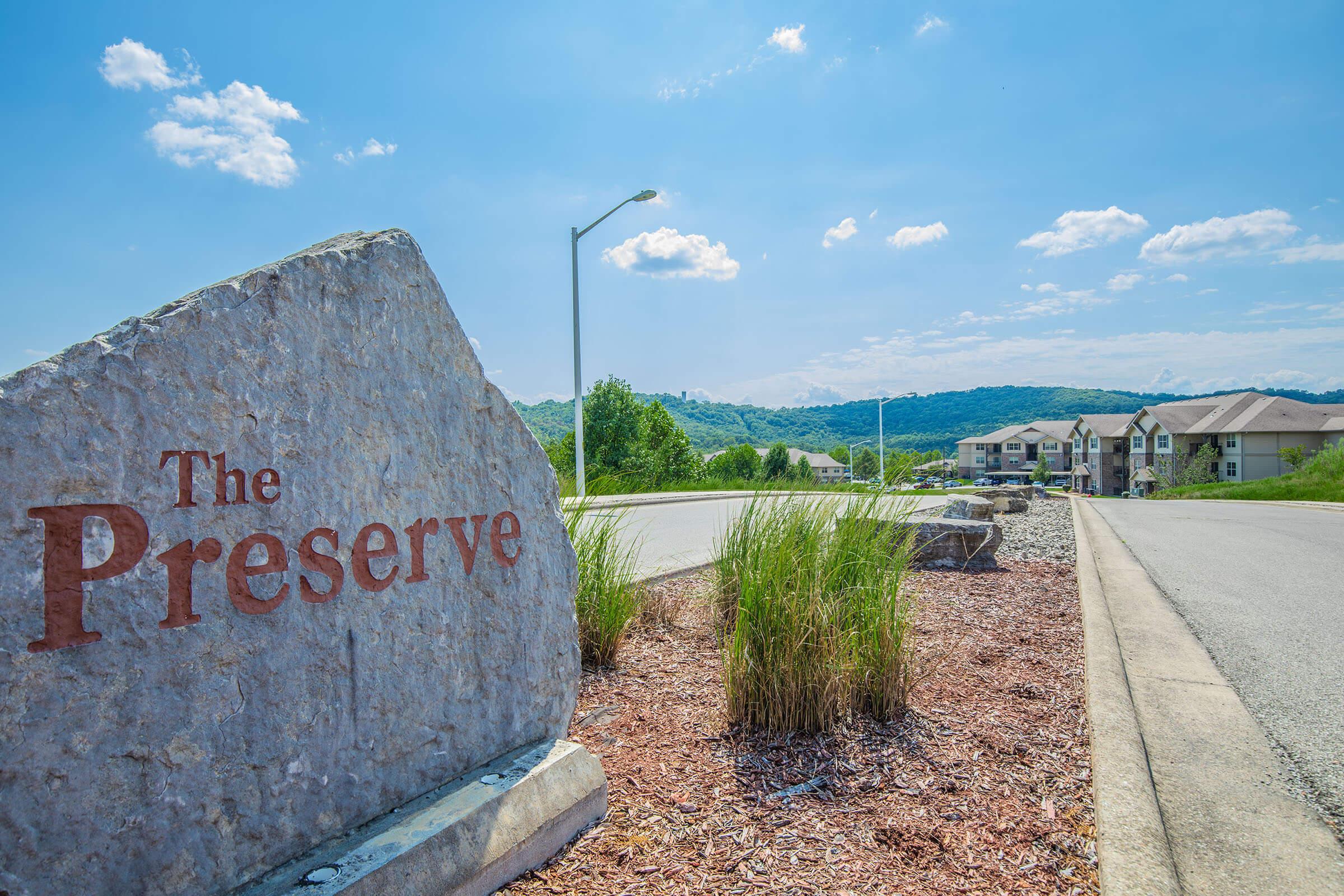 A large gray stone with "The Preserve" engraved on it stands at the entrance to a residential area. In the background, green hills rise under a bright blue sky dotted with fluffy white clouds. Apartment buildings are visible along the road, surrounded by landscaped greenery.