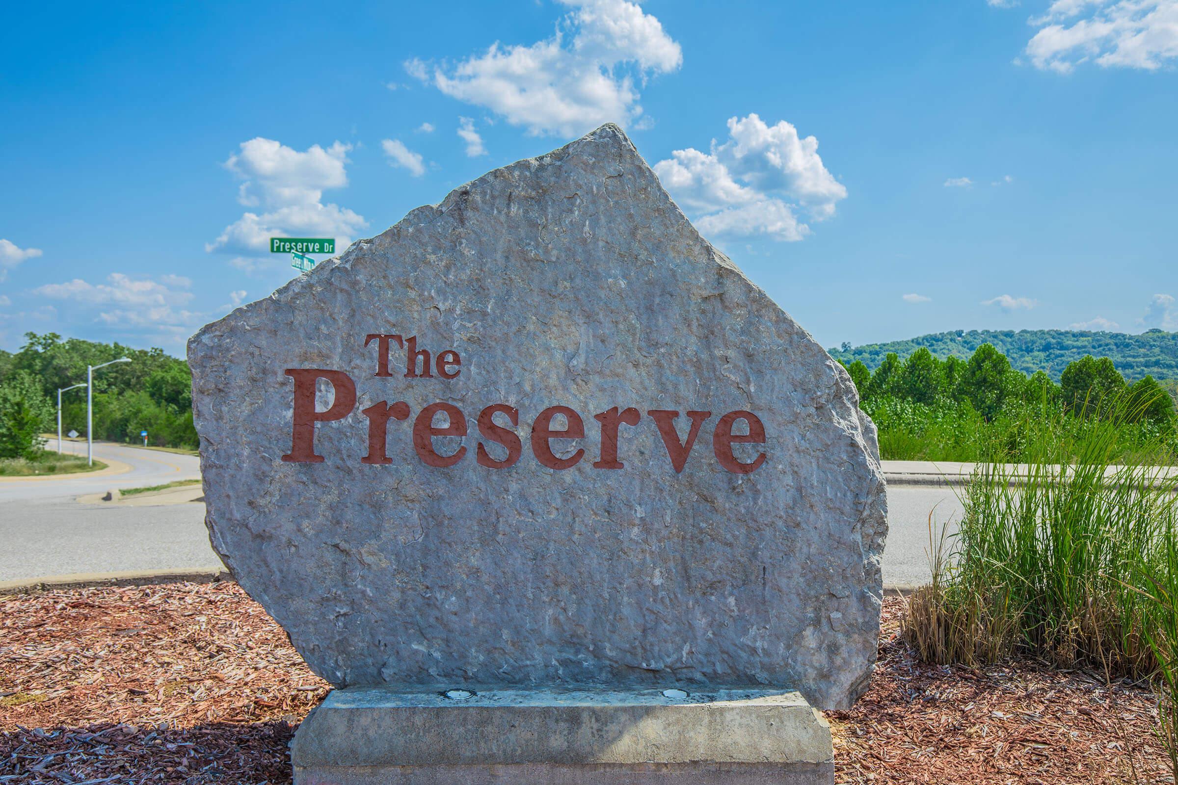 Large stone marker with the engraved text "The Preserve." It is set against a bright blue sky with fluffy white clouds. The surrounding landscape includes grassy areas and trees in the background, indicating a natural setting. A road sign for "Preserve" is visible in the distance.