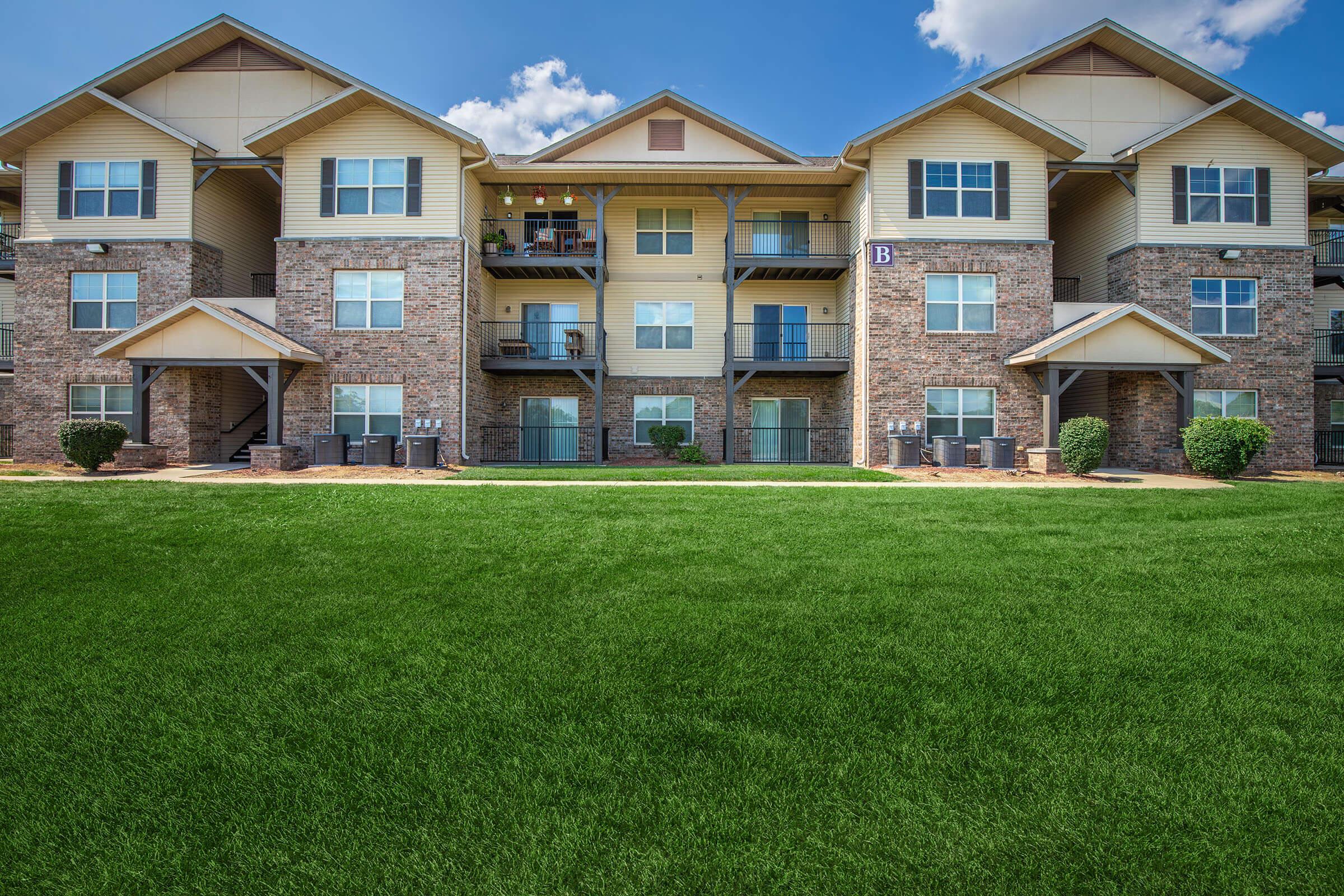 A modern apartment building featuring multiple levels with balconies, surrounded by well-maintained green grass. The facade includes a mix of brick and siding, under a clear blue sky with a few clouds. The scene conveys a welcoming residential atmosphere.