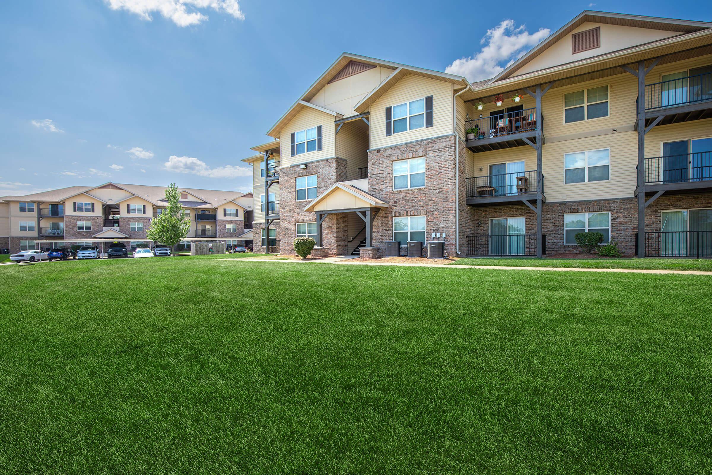 A sunny view of a multi-story apartment building with balconies and landscaped green lawns. Cars are parked nearby, and the sky is clear with a few clouds, creating a pleasant residential atmosphere.