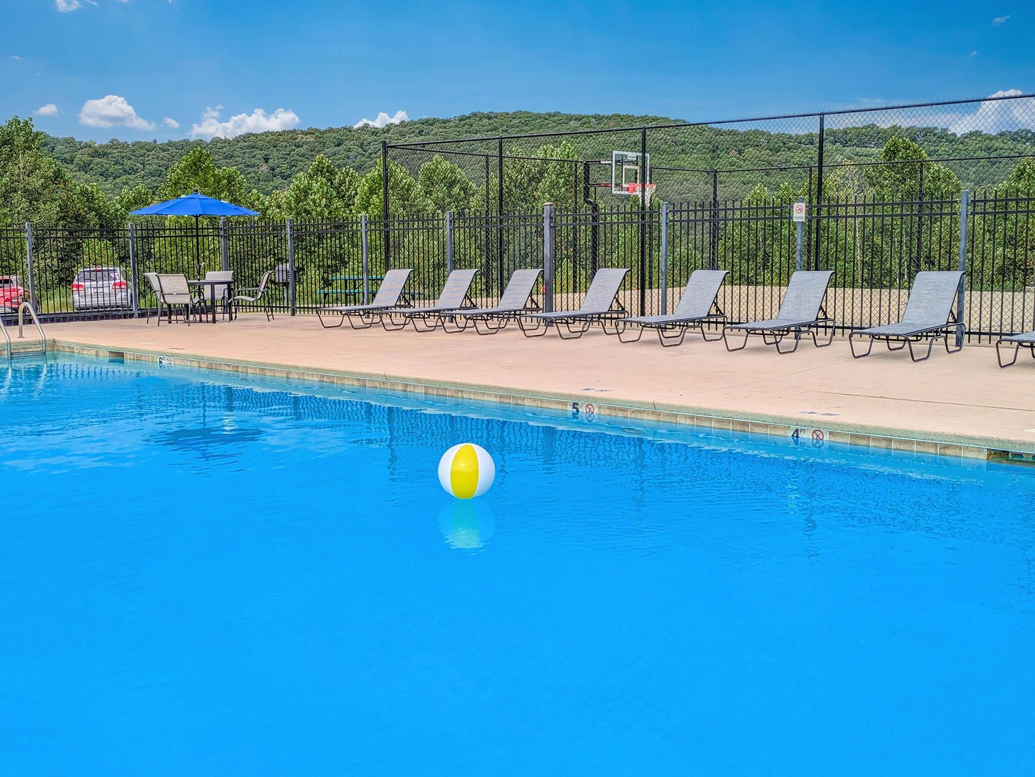 A serene outdoor pool surrounded by lounge chairs and a colorful beach ball floating in the water. In the background, green hills are visible under a clear blue sky, with a basketball hoop near the pool area. The setting is perfect for relaxation and recreation.