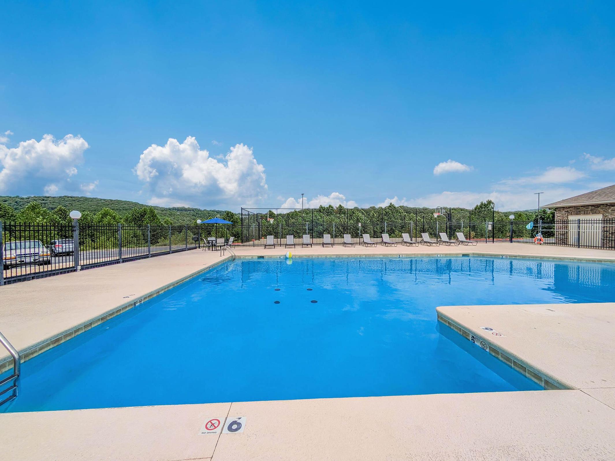 A bright blue swimming pool surrounded by a fenced area, with several lounge chairs lined up on one side. The sky is clear with a few fluffy clouds, and green hills are visible in the background. A few umbrellas are set up nearby, indicating a recreational area for relaxation and enjoyment.