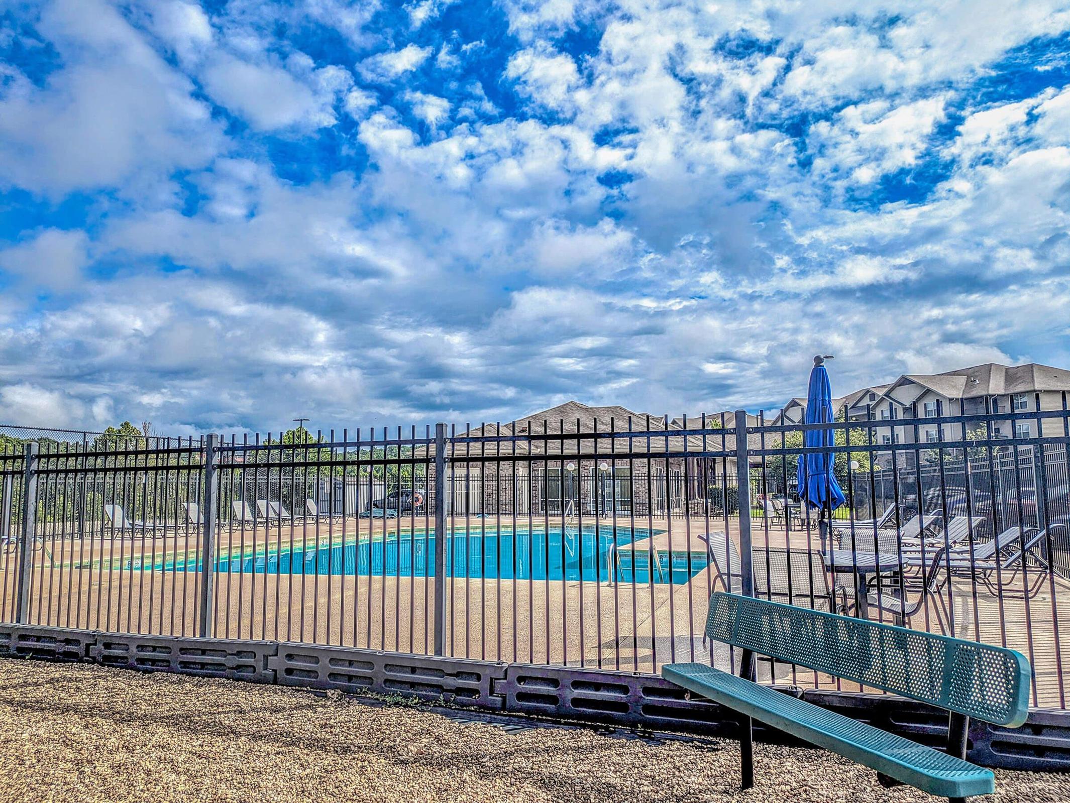 A blue swimming pool surrounded by a black metal fence, with lounge chairs neatly arranged around it. In the background, there are two-story buildings. The sky is partly cloudy with patches of blue, creating a serene atmosphere. A green bench is placed in the foreground on a gravel surface.