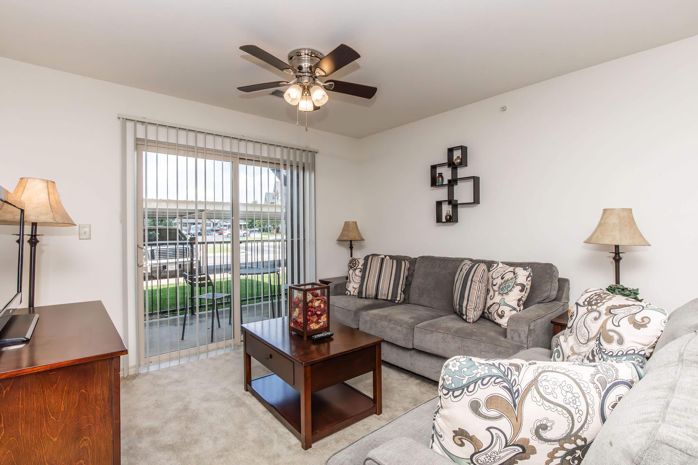 Cozy living room featuring a gray couch with decorative pillows, a wooden coffee table with a centerpiece, and two floor lamps. A ceiling fan hangs above, and large sliding glass doors provide access to a patio area. Neutral-colored walls create a warm and inviting atmosphere.