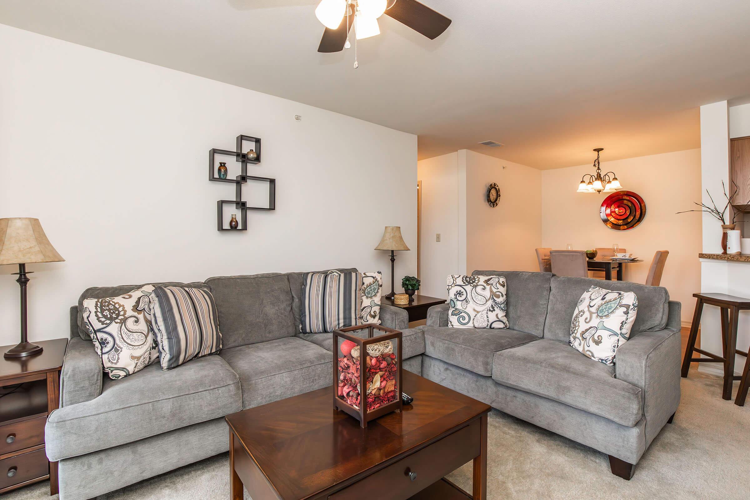A cozy living room featuring a gray sectional sofa with decorative pillows, a wooden coffee table with a floral centerpiece, and side lamps. In the background, there is a dining area with a round table, chandelier, and wall art, creating a welcoming and stylish atmosphere.