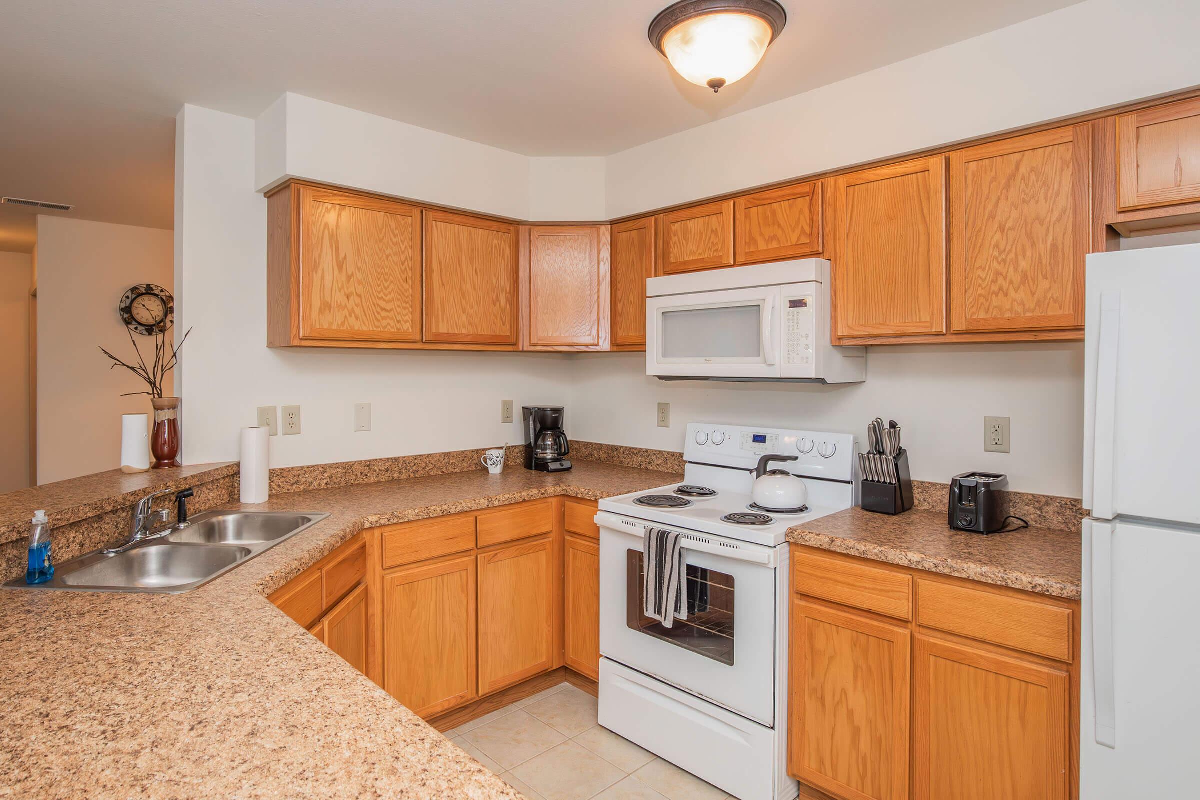 A modern kitchen featuring wooden cabinets, a white microwave and oven, and a double sink. The countertops are made of speckled brown granite, and there's a coffee maker and a toaster visible. Natural light illuminates the space, enhancing the warm tones of the cabinetry.