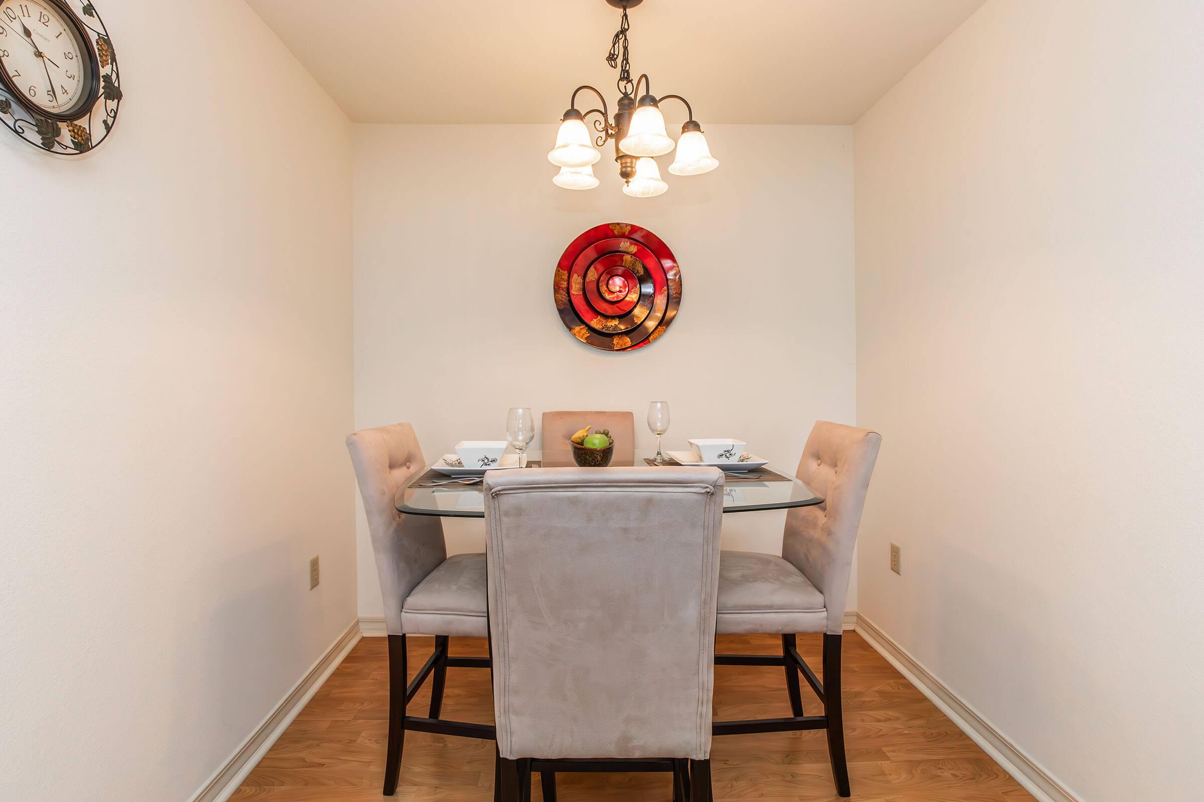A cozy dining area featuring a round glass table with two place settings. There are four upholstered chairs, a decorative fruit bowl at the center, and a colorful circular wall art piece. Soft lighting from a chandelier above creates a warm atmosphere, with a wall clock visible on the left.