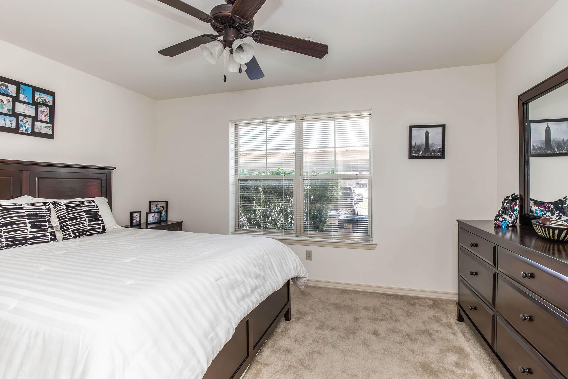 A cozy bedroom featuring a king-size bed with white and black striped bedding, a dark wood dresser, and framed photographs on the walls. A large window allows natural light to enter, with views of green shrubbery outdoors. There’s a ceiling fan for added comfort.