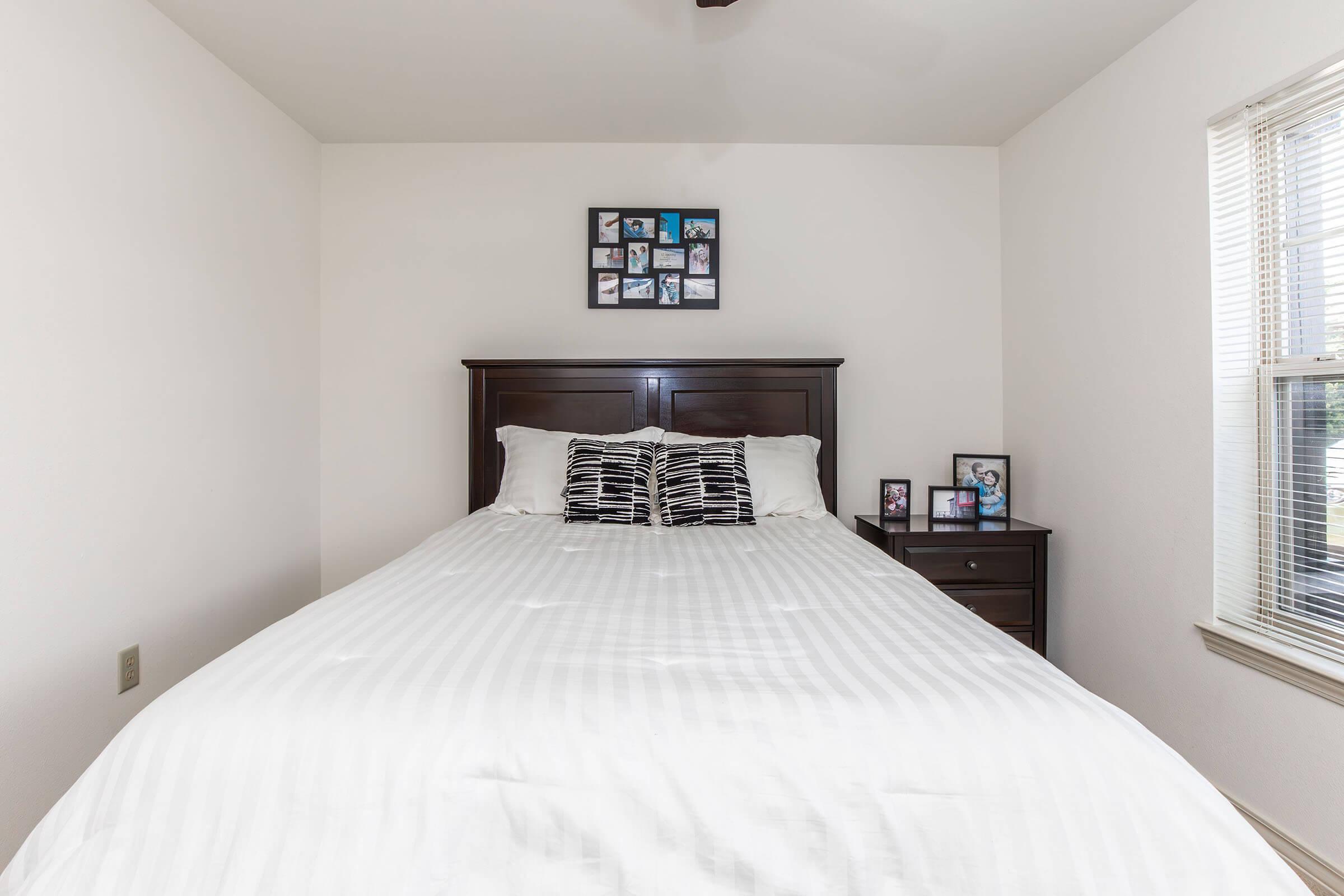 A neatly made bed with white bedding featuring black and white striped pillows, positioned against a plain wall. To the right, a bedside table holds small framed photographs. A window with blinds allows natural light into the room, creating a bright and inviting atmosphere.