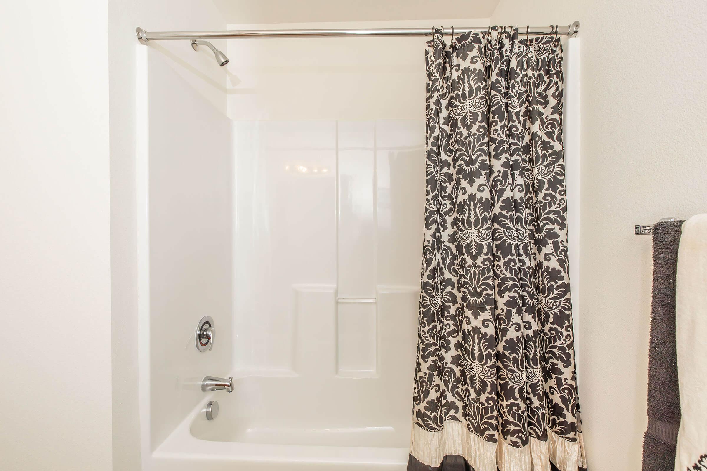A clean, modern bathroom featuring a white bathtub with a shower. The shower area is partially covered by an elegant black and white patterned curtain. A towel hangs on a bar nearby, complementing the aesthetic with neutral tones. The walls are white, creating a bright, airy atmosphere.