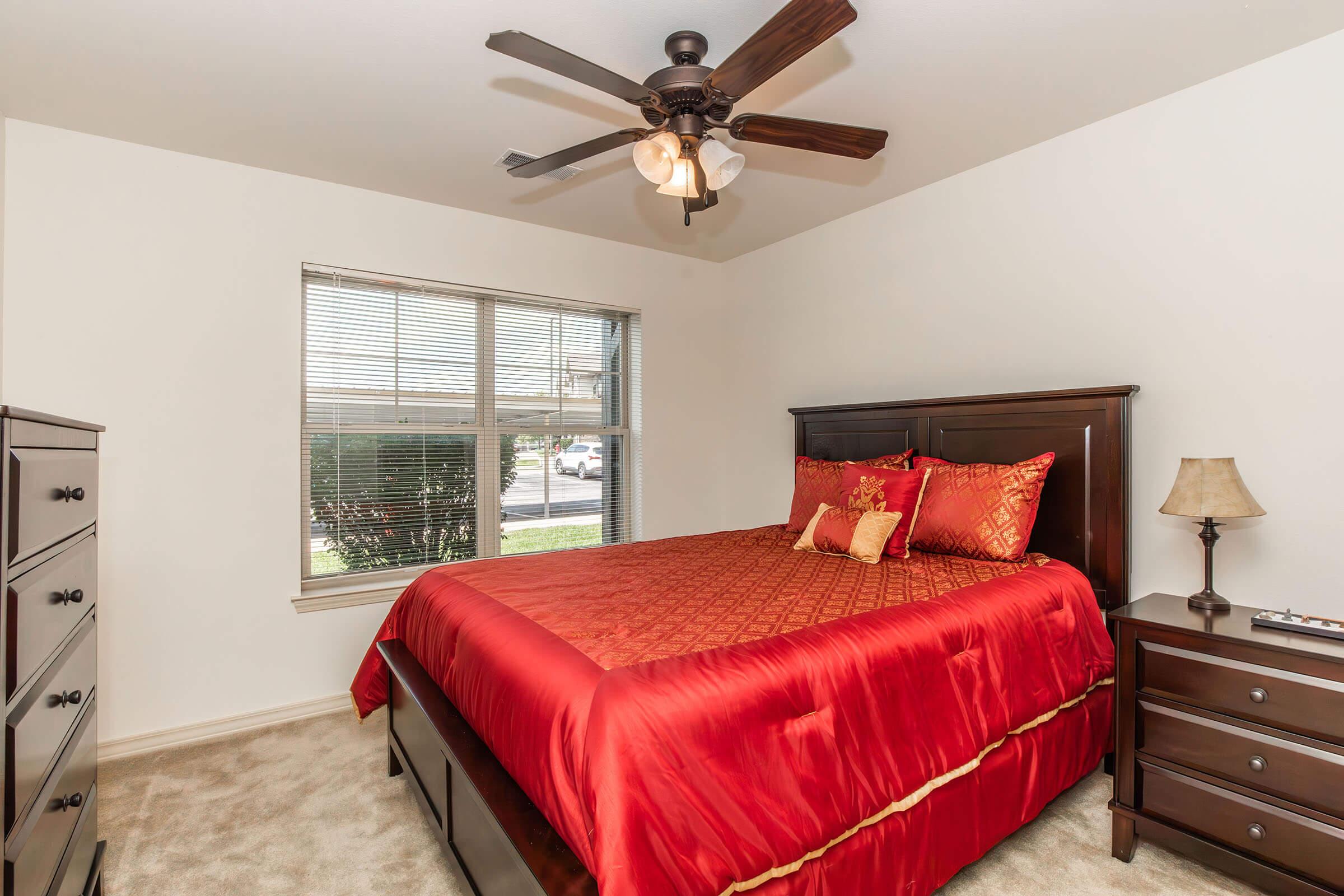 A cozy bedroom featuring a wooden bed with a bright red comforter and matching pillows. It includes a bedside lamp and a dresser, with a ceiling fan above. A window lets in natural light, revealing a view of greenery outside. The walls are painted white, creating a bright and inviting atmosphere.