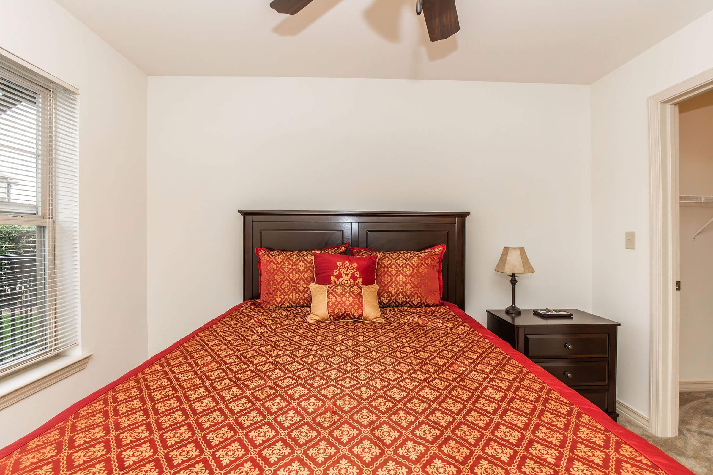 A cozy bedroom featuring a dark wood bed frame with a decorative red and gold patterned bedspread and matching pillows. A bedside table with a lamp is positioned next to the bed, and a window with blinds allows natural light to fill the room. A closet door is visible in the background.