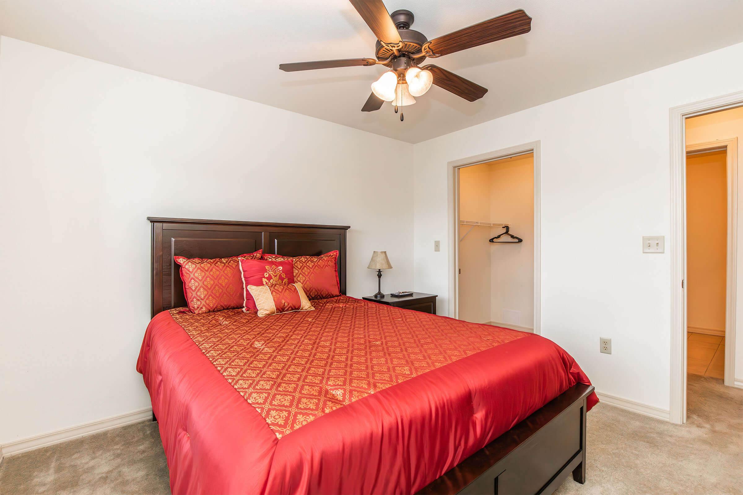 A cozy bedroom featuring a dark wooden bed with a red patterned comforter and matching pillows. A bedside lamp and a ceiling fan enhance the decor. A closet space is visible in the background, and the walls are painted white, creating a bright and inviting atmosphere.
