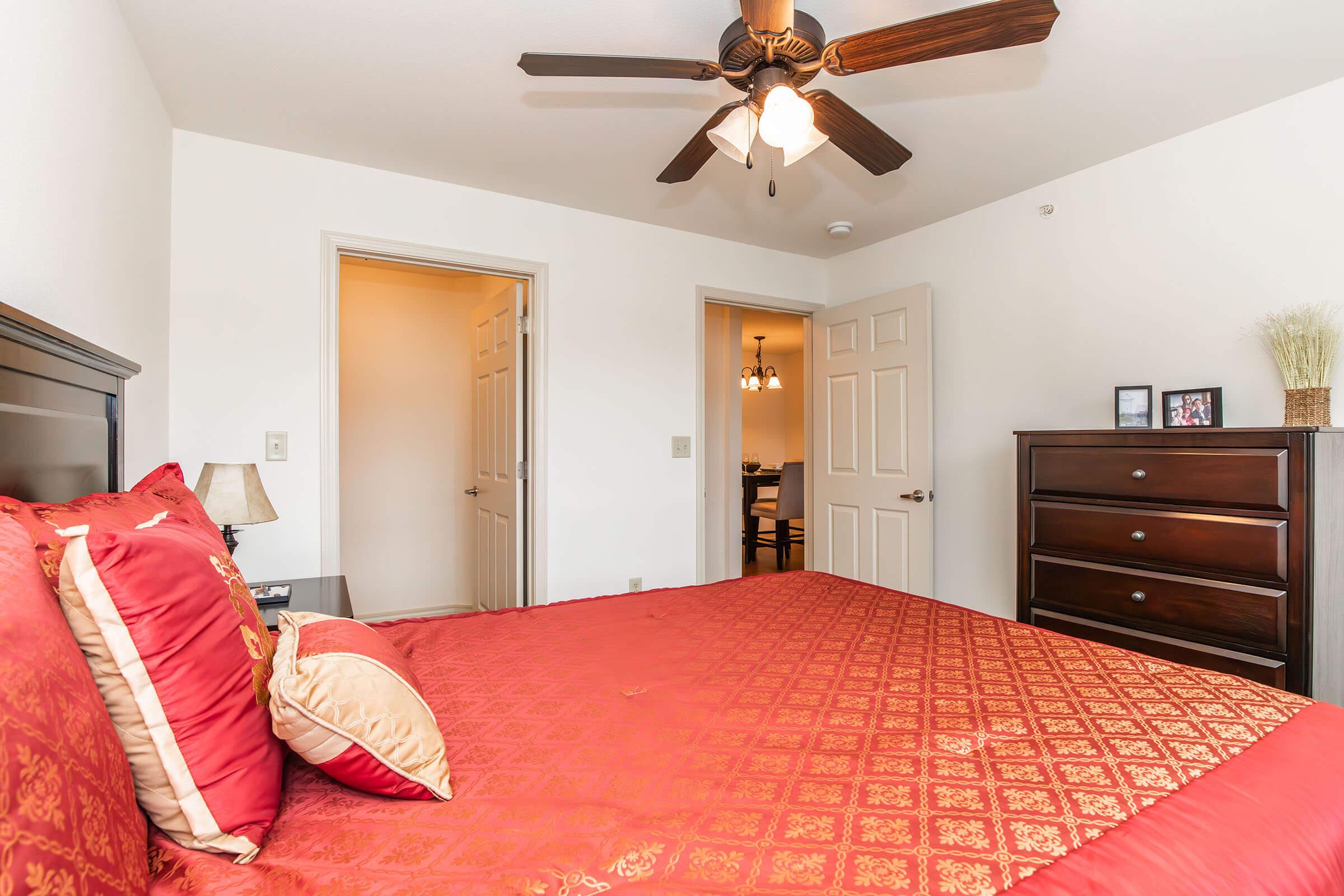 A well-decorated bedroom featuring a large red bedspread with decorative pillows, a wooden dresser, and a ceiling fan. The room includes open doors leading to a hallway and adjacent areas, creating a bright and inviting atmosphere with light-colored walls and minimal decor.