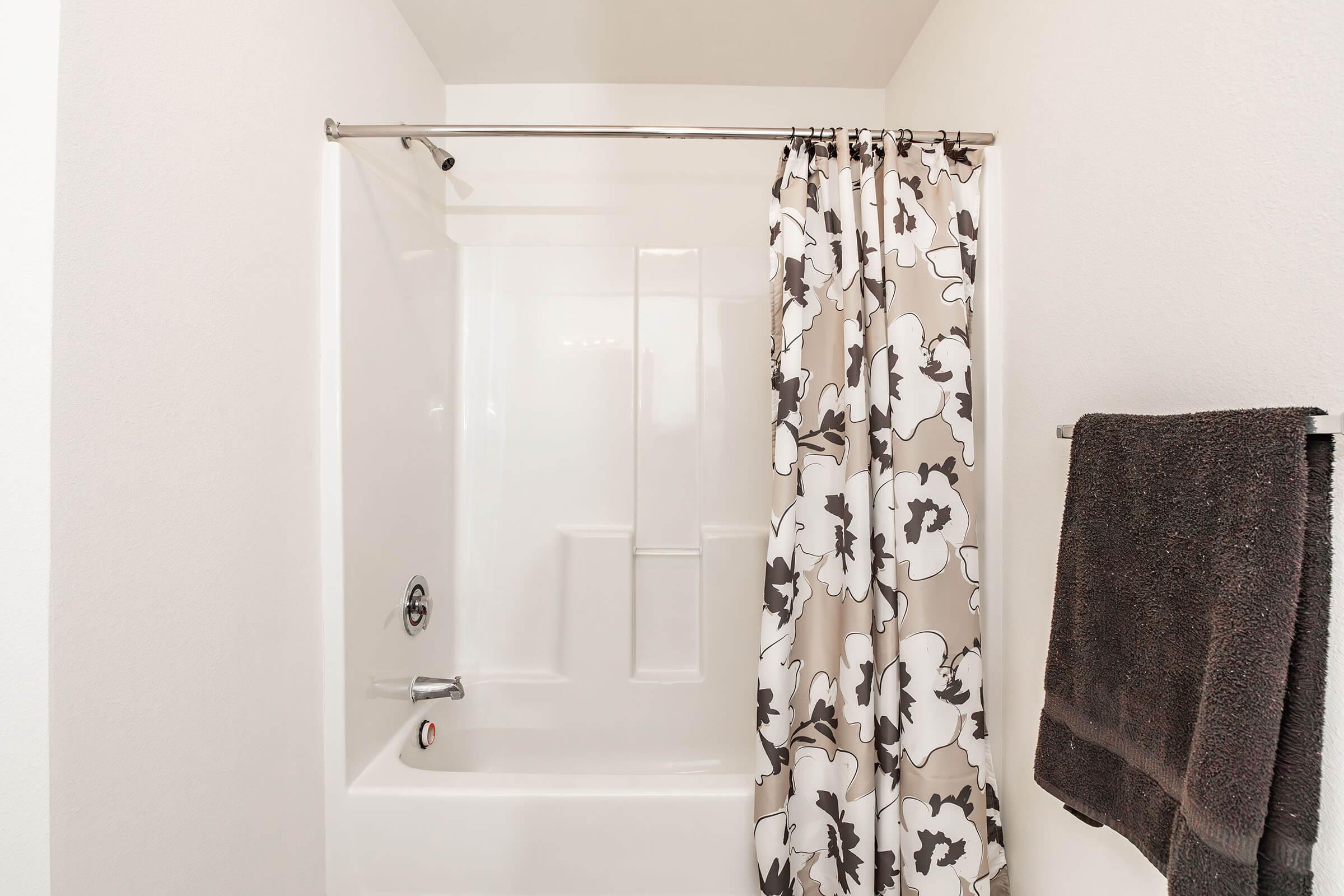 A clean bathroom with a white bathtub and shower combination. The shower has a patterned curtain featuring large flowers in neutral tones, and a dark gray towel hangs on the wall nearby. The overall atmosphere is bright and minimalist.
