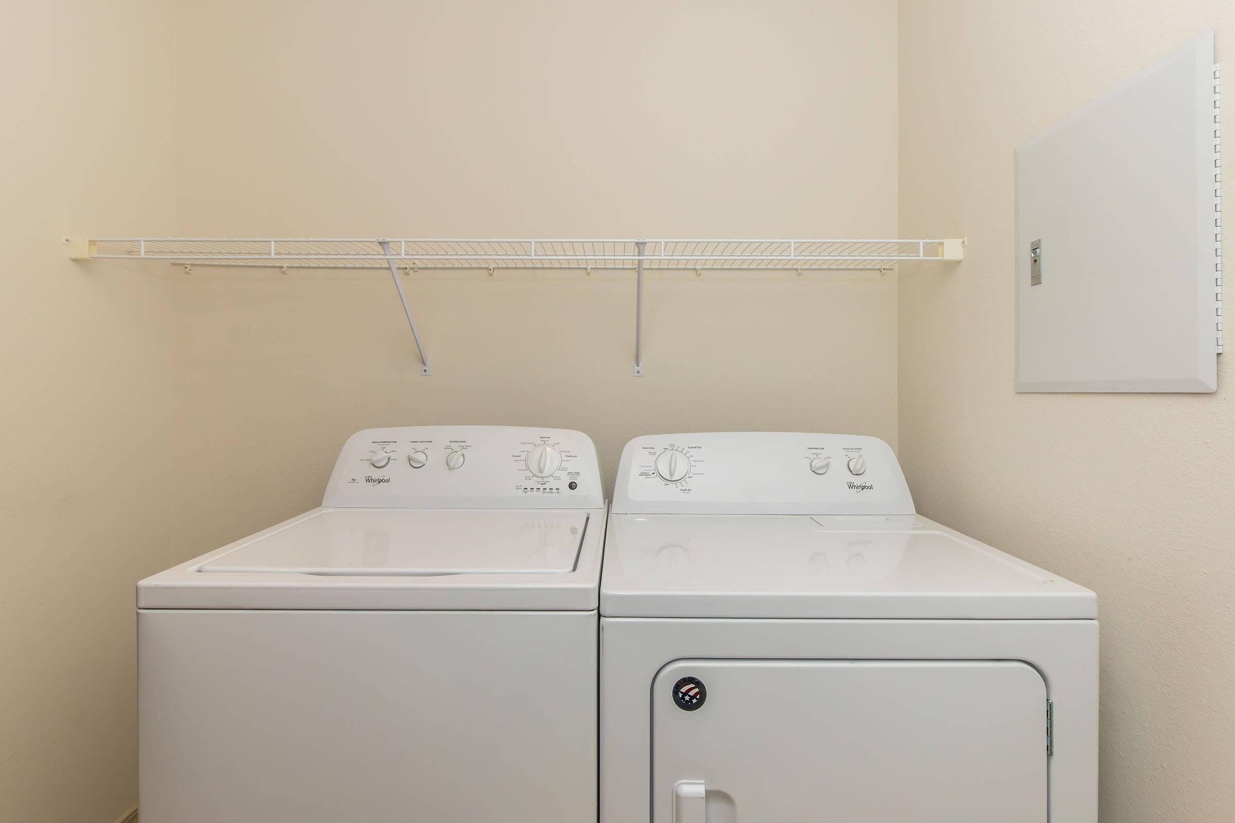 A clean, organized laundry room featuring a white washer and dryer side by side. Above them, there is a wire shelf for storage. The walls are a light beige color, providing a bright and tidy environment.