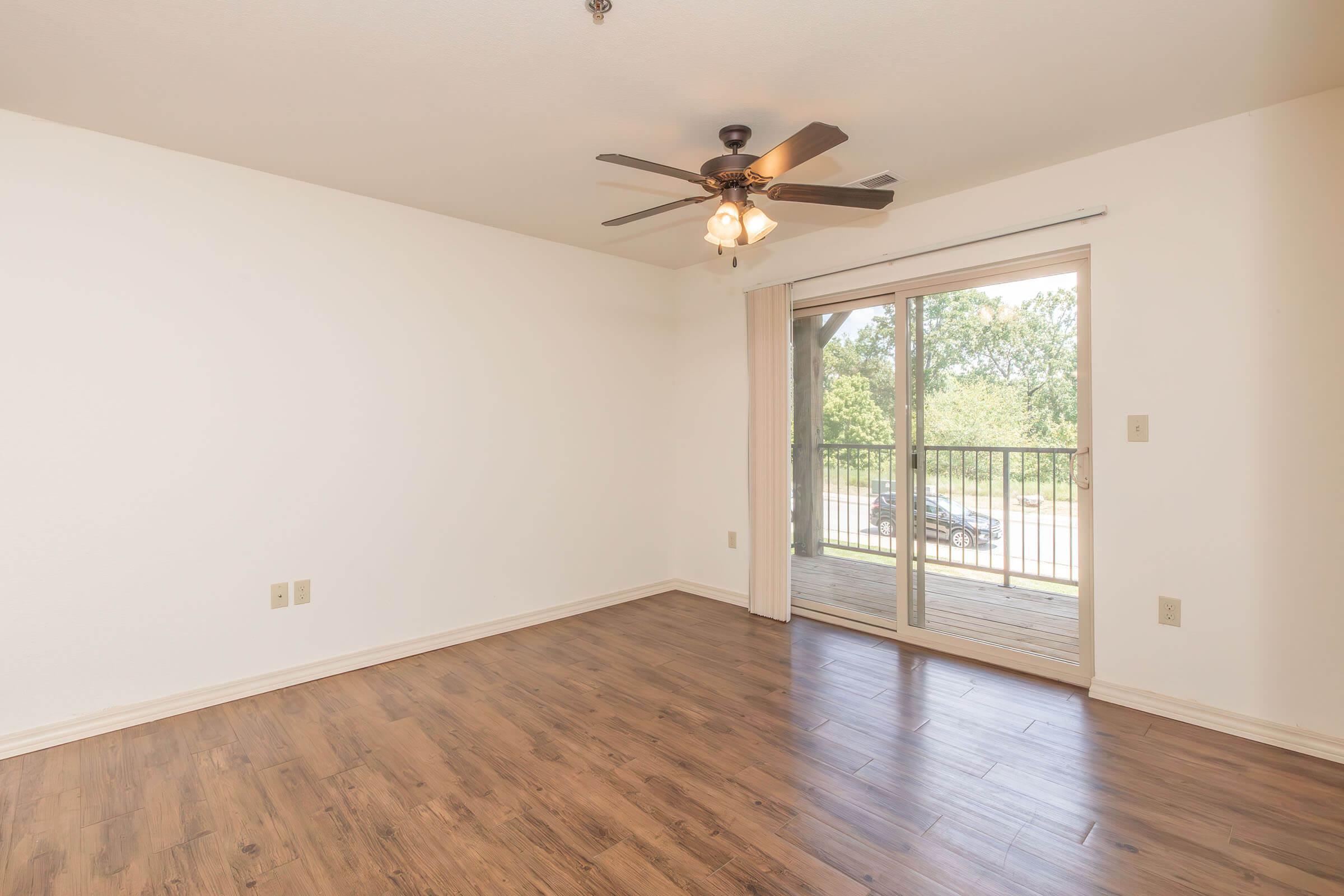 Empty room with light-colored walls and a ceiling fan. Large sliding glass door leads to a balcony, allowing natural light to enter. Wood-like flooring covers the ground, creating a warm atmosphere. The space is uncluttered, ideal for personalized decoration.
