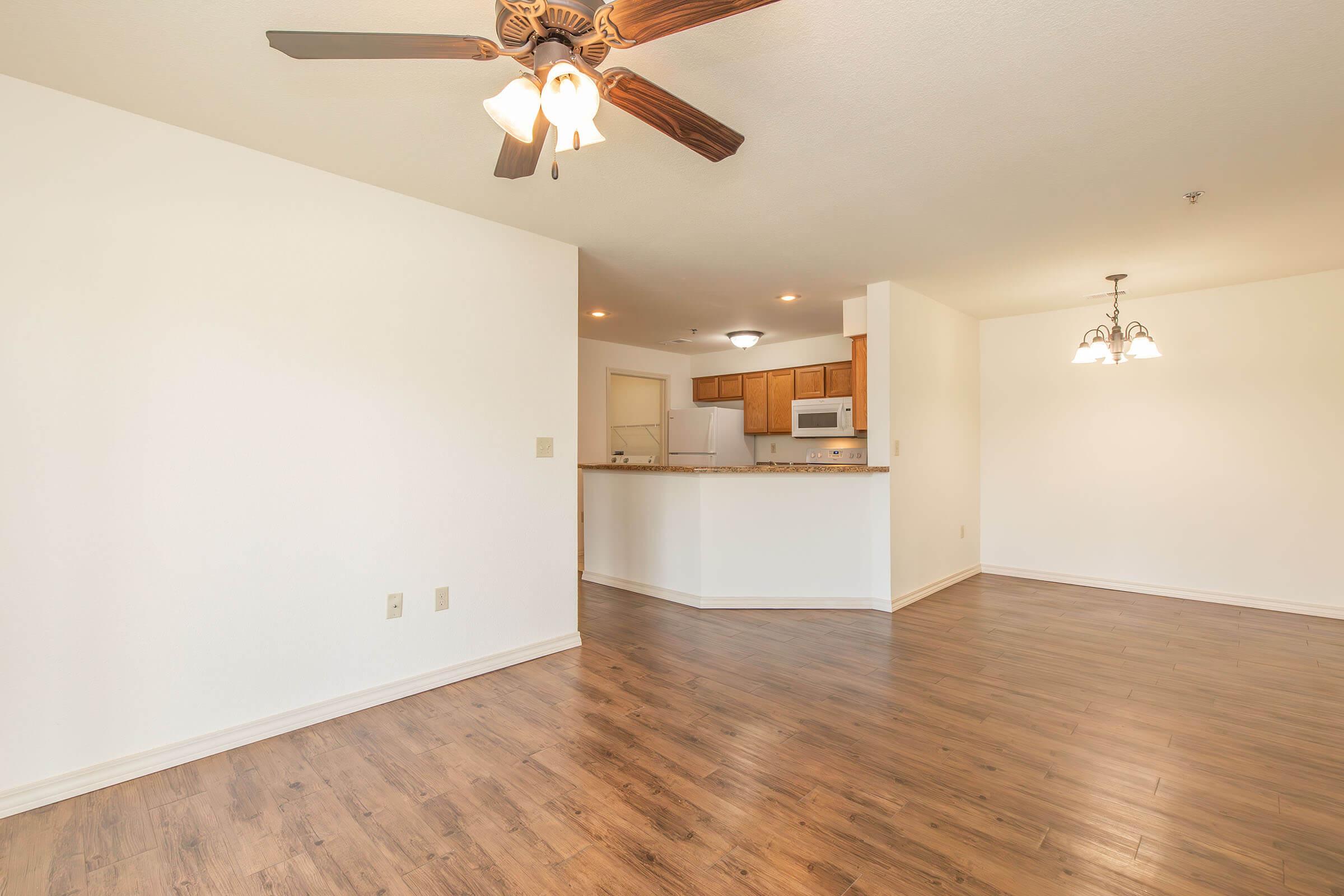 A bright and spacious living area with wooden flooring, featuring a ceiling fan and light fixture. The room has an open layout that leads to a kitchen with white cabinets and appliances. Natural light enters through the windows, creating a warm and inviting atmosphere.