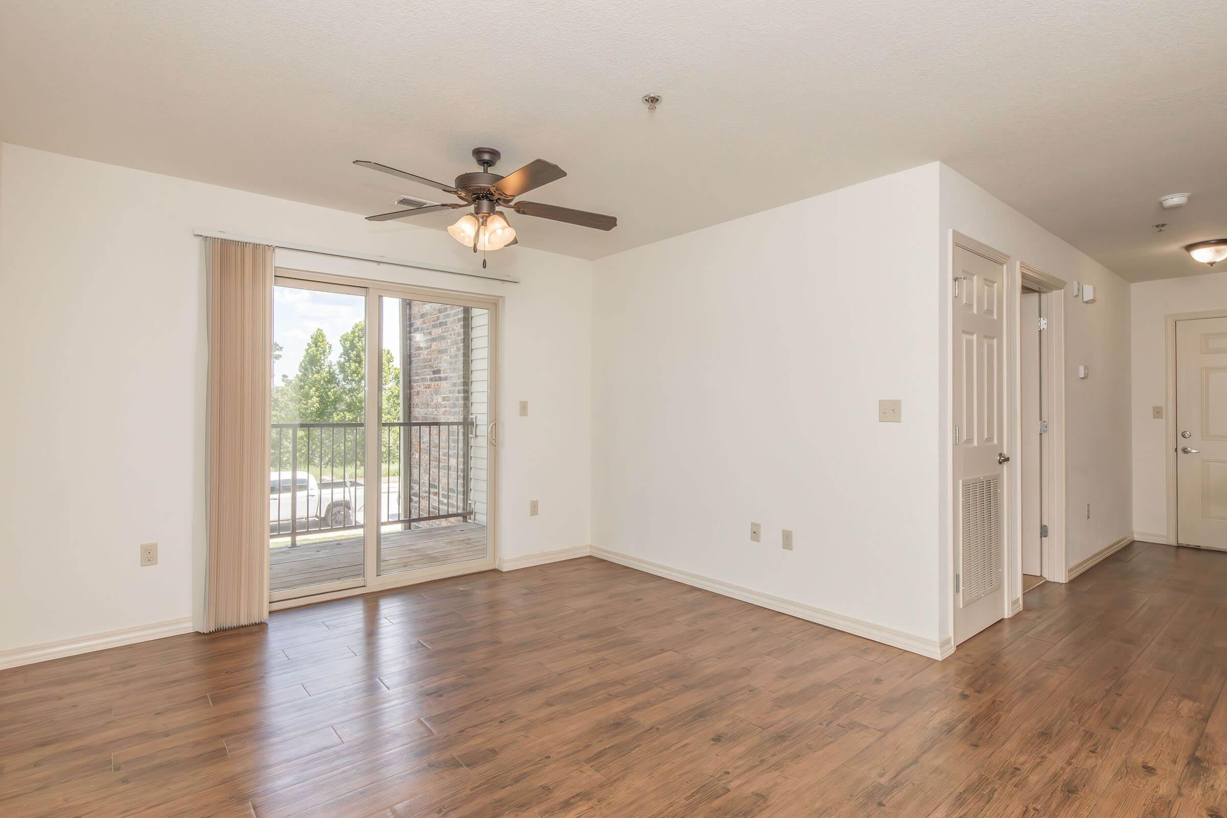 A spacious room featuring a ceiling fan, hardwood flooring, and beige walls. There is a large sliding glass door leading to a balcony, providing natural light. Doorways to other rooms are visible, adding to the open layout of the living space.
