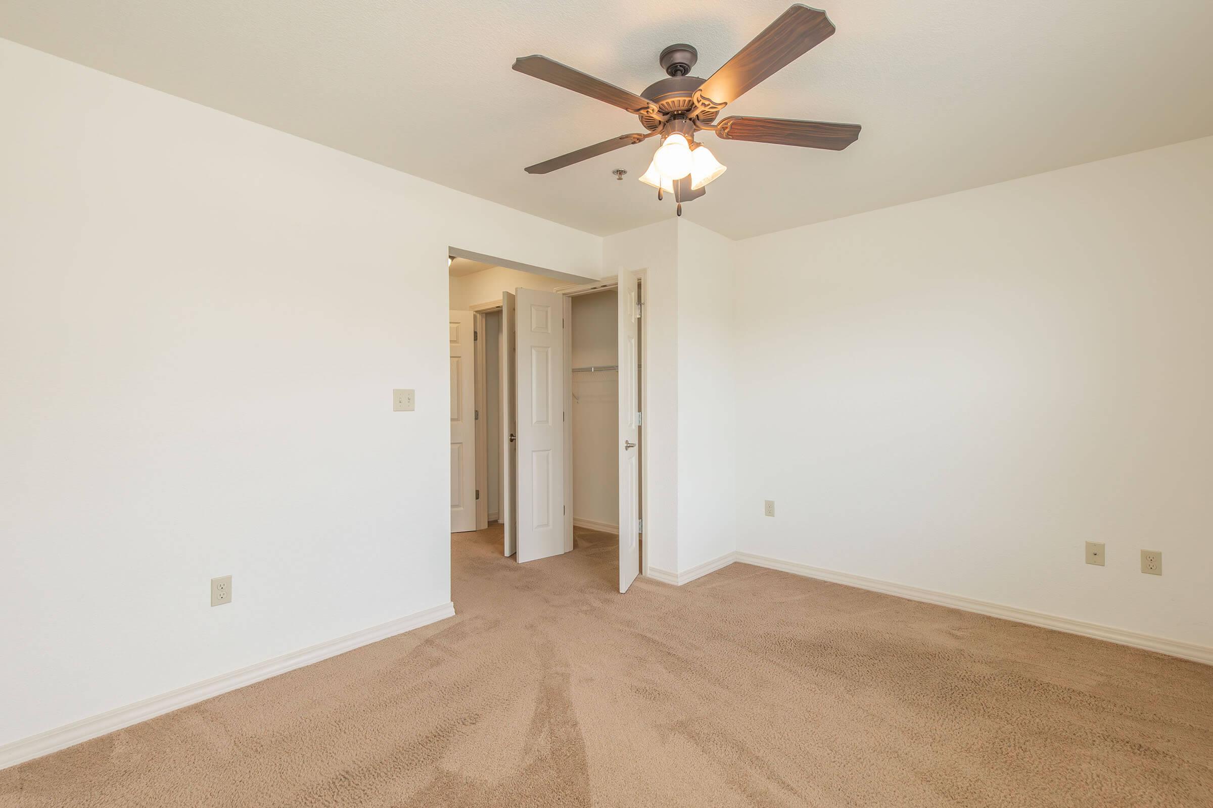 A spacious, well-lit bedroom featuring a ceiling fan with light, neutral-colored walls, and plush carpeting. The room has a doorway leading to a closet and another entrance, creating an inviting and airy atmosphere.